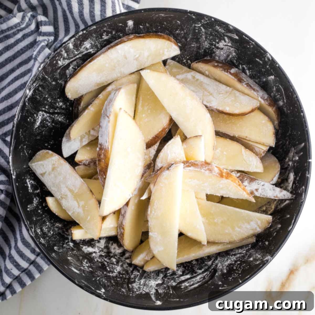 Potato wedges in a dark bowl, tossed with white arrowroot powder. 