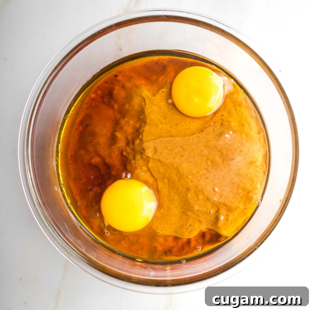A person mixing wet ingredients (nut butter, maple syrup, eggs/flax eggs) in a medium glass bowl with a spatula, creating a thick, uniform mixture.