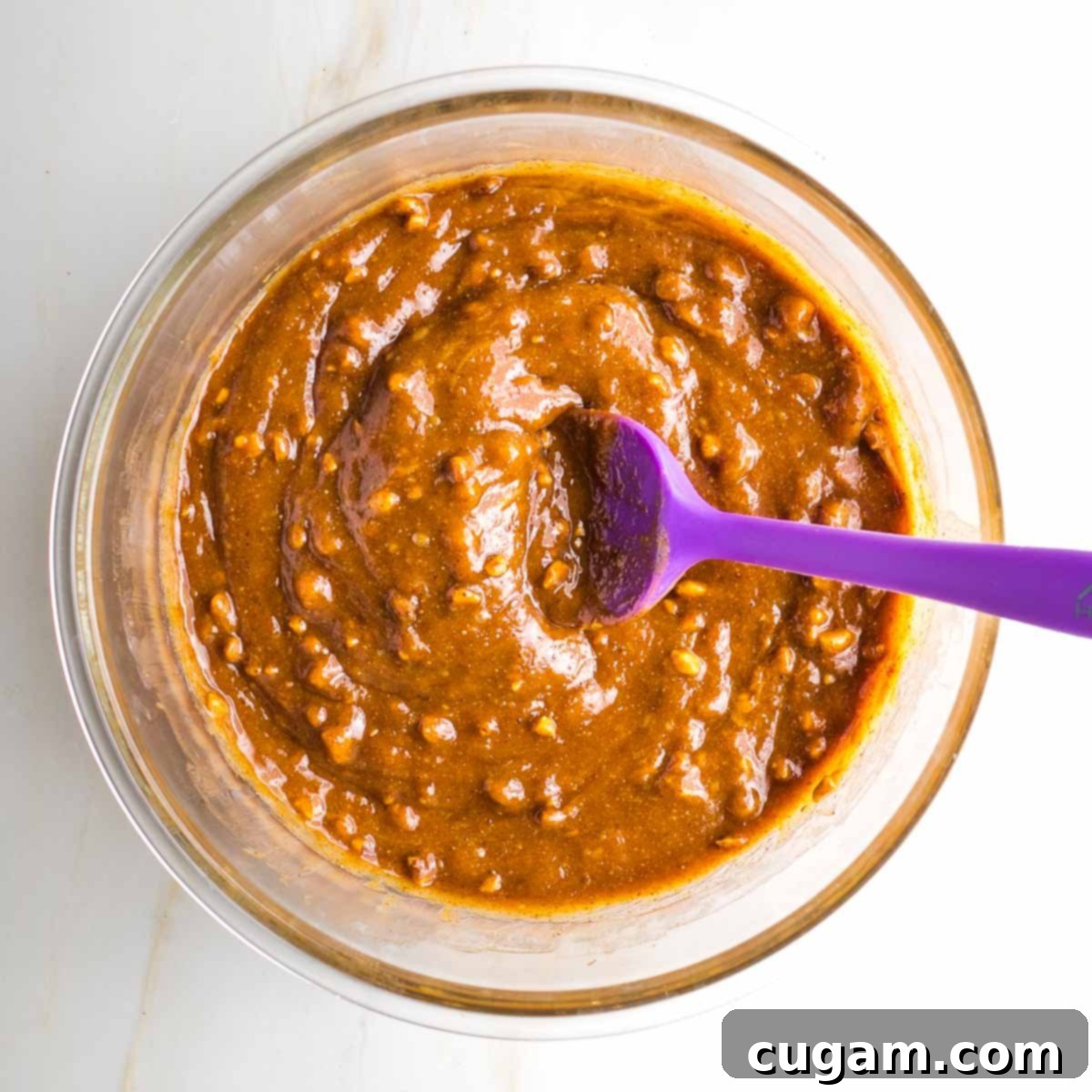 Close-up of thick, sticky cookie dough being mixed in a glass bowl with a rubber spatula, showing the lumpy texture from crunchy almond butter.