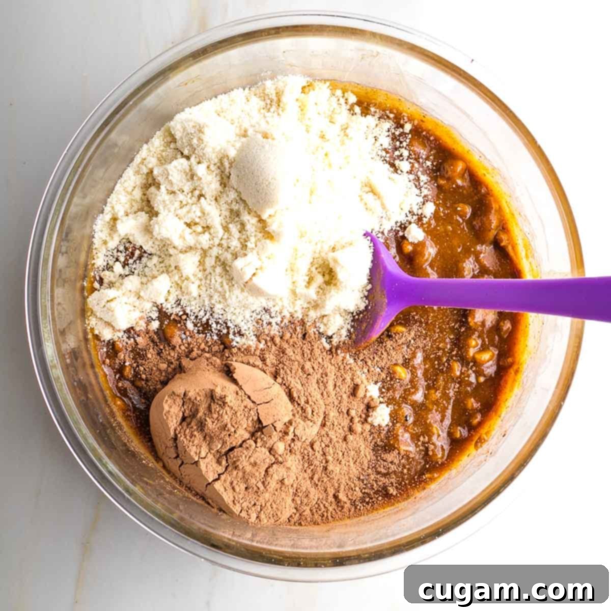 Almond flour and cacao powder sprinkled over the wet cookie dough ingredients in a glass bowl.