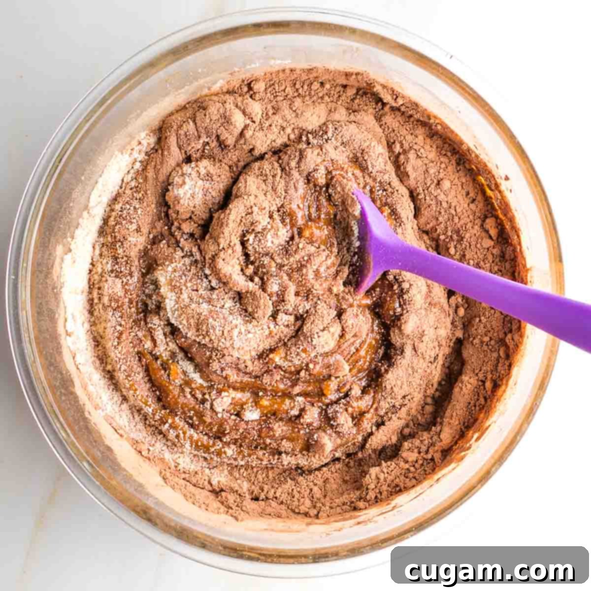 Mixing chocolate cookie dough in a glass bowl with a purple spatula, ensuring no dry streaks remain.