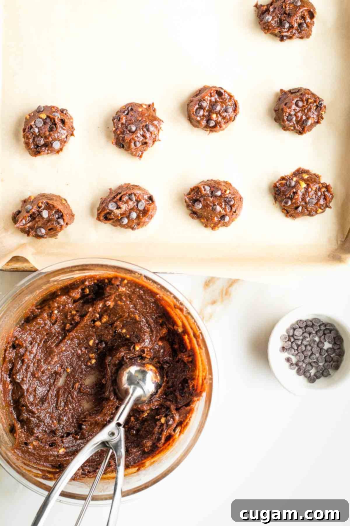 A person using a silver cookie scoop to neatly portion out sticky chocolate cookie dough onto a baking sheet lined with parchment paper.
