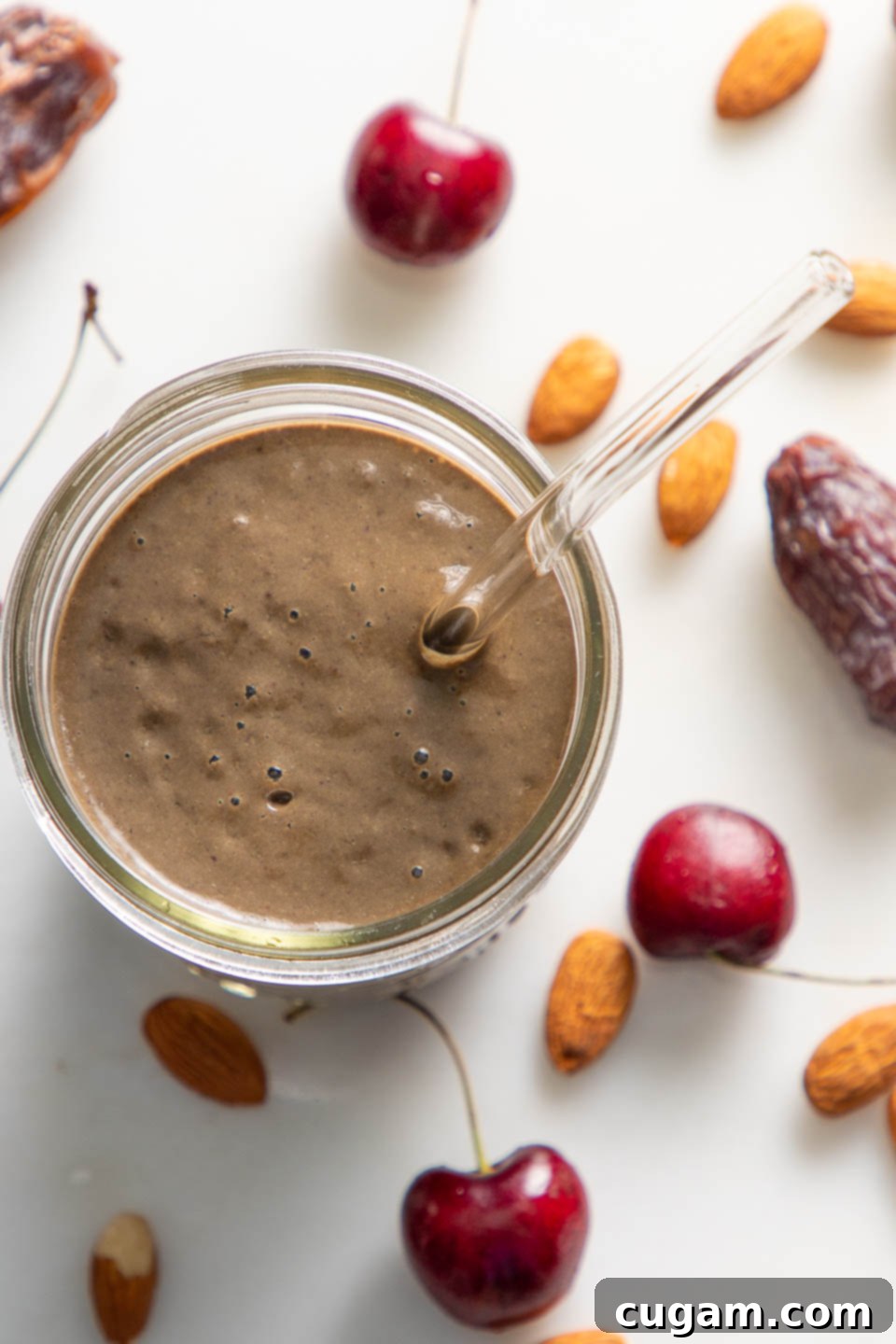 Cocoa Cherry Indulgence 5 Chocolate smoothie in a glass with straw. Countertop has cherries, almonds and dates.