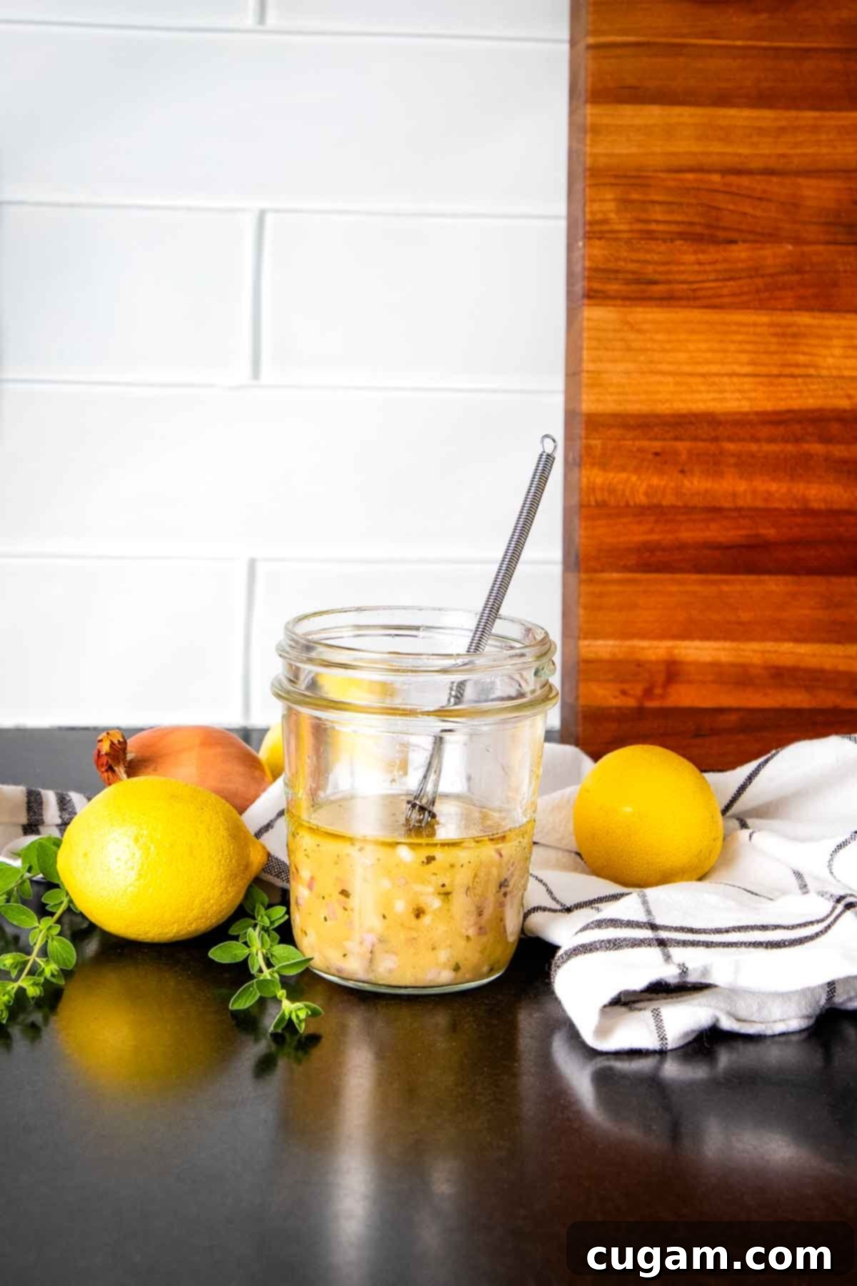 A jar of freshly made yellowish salad dressing with a small whisk, surrounded by fresh lemons and herbs on a kitchen counter.