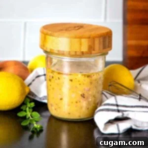 Glass jar with wooden lid filled with homemade lemon vinaigrette, surrounded by fresh lemons, a shallot, and some fresh herbs on a light surface.