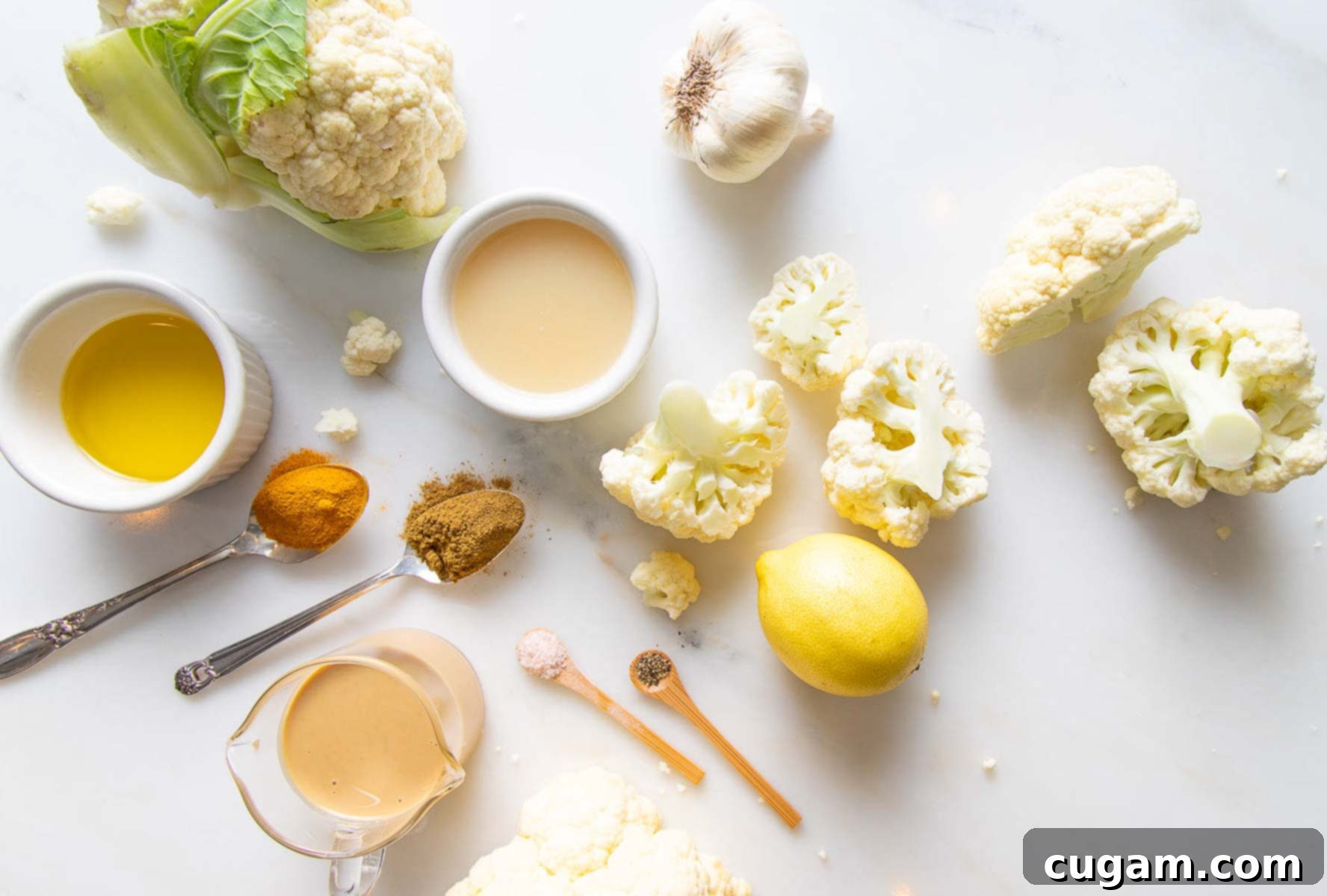 Ingredients for roasted cauliflower hummus laid out on a table: cauliflower, garlic, tahini, lemon, spices, olive oil.