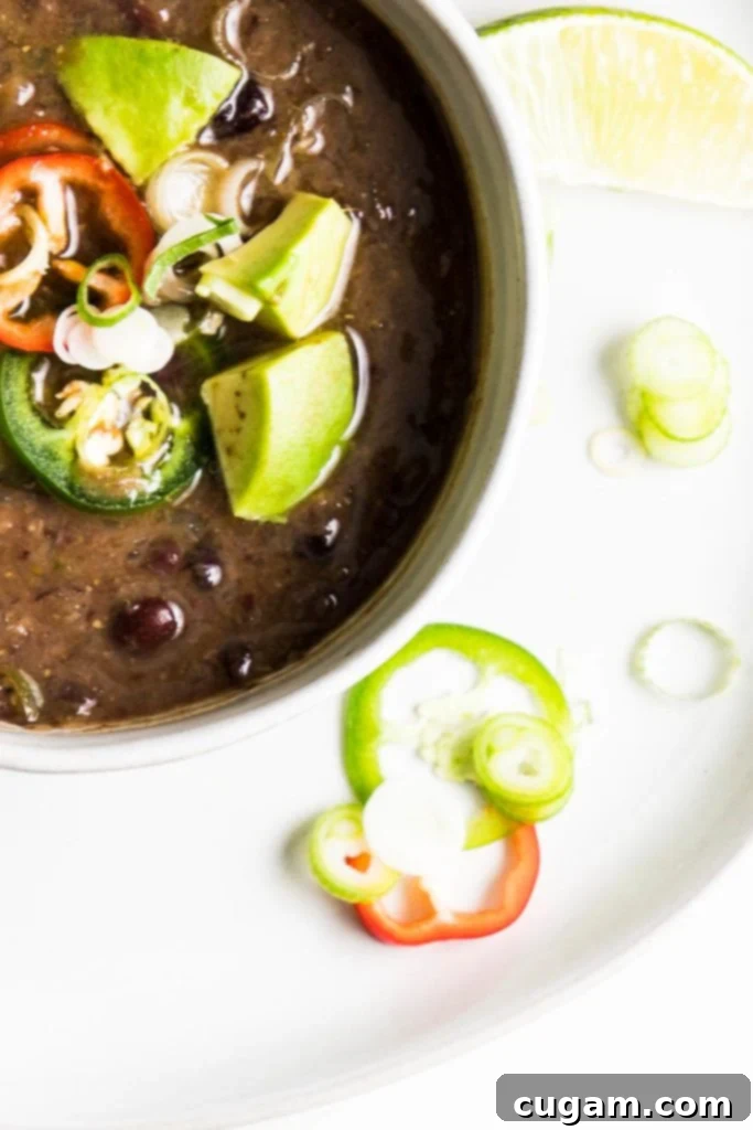 Vegan black bean soup in a white bowl, generously topped with fresh avocado chunks, sliced jalapeños, a lime wedge, and chopped scallions.