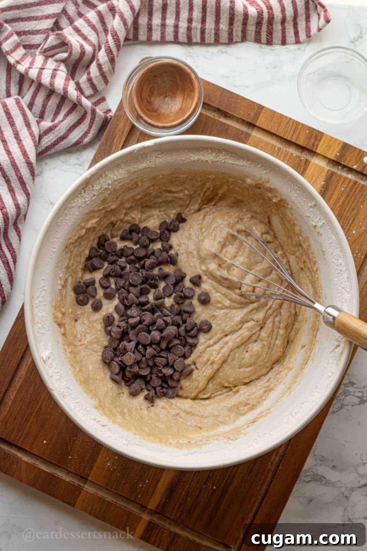 Elevated Sourdough Banana Bread 8 Chocolate chips being stirred into the raw banana bread batter in a white bowl with a whisk.
