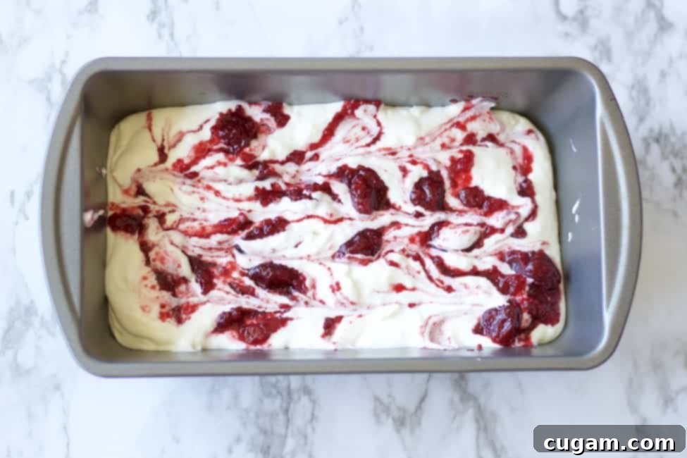 No Churn Cherry Cheesecake Ice cream with graham cracker crumbles, vanilla, sweetened condensed milk, cream cheese, and cherry pie filling. Made with real cream because nothing else will do. Best summer dessert ever! #icecream #nochurnicecream #easydessert #cherrycheesecake #cherrydesserts #cheesecakedessert #fruitfilleddessert #summertreats Rich cherry pie filling being swirled artfully into a pan of creamy whipped cream, creating beautiful patterns for the ice cream.
