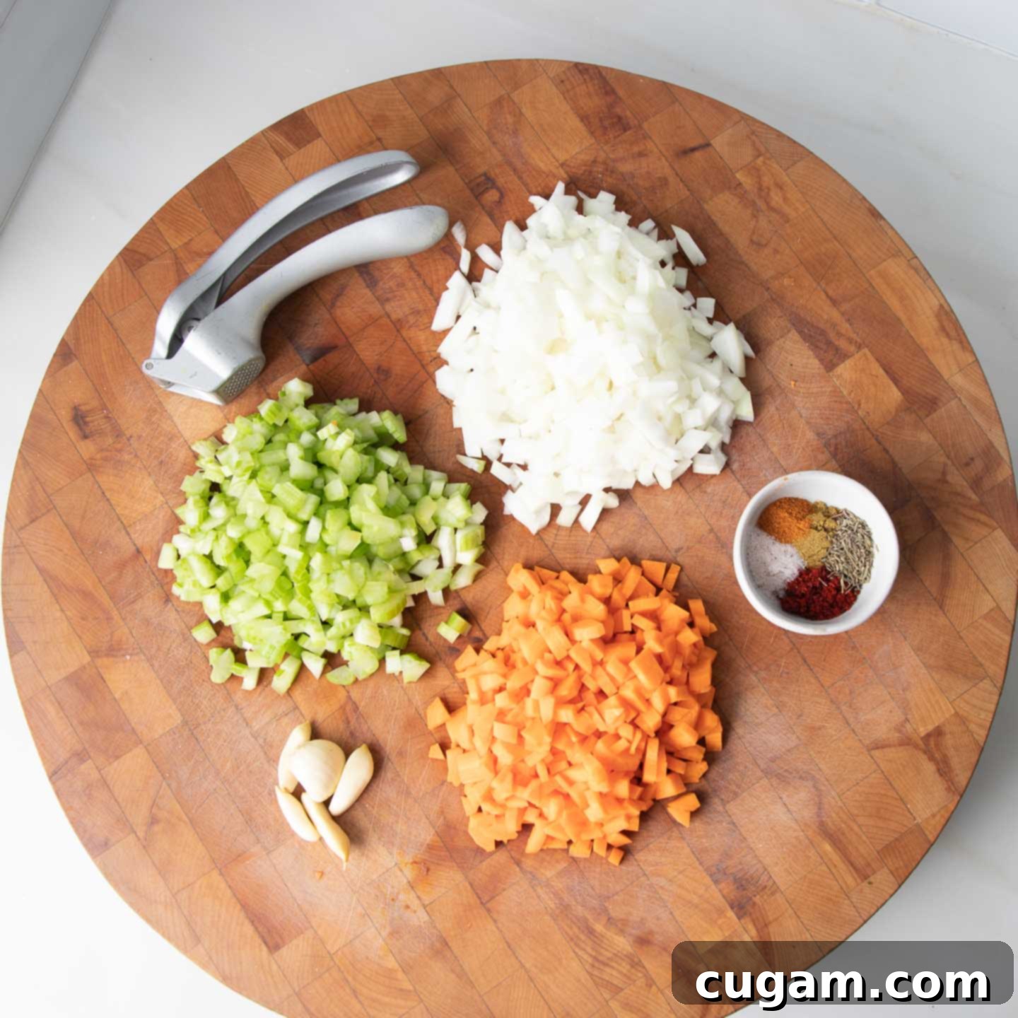 A round wooden board displaying meticulously diced onions, celery, carrots, and garlic, alongside a small dish of measured spices.