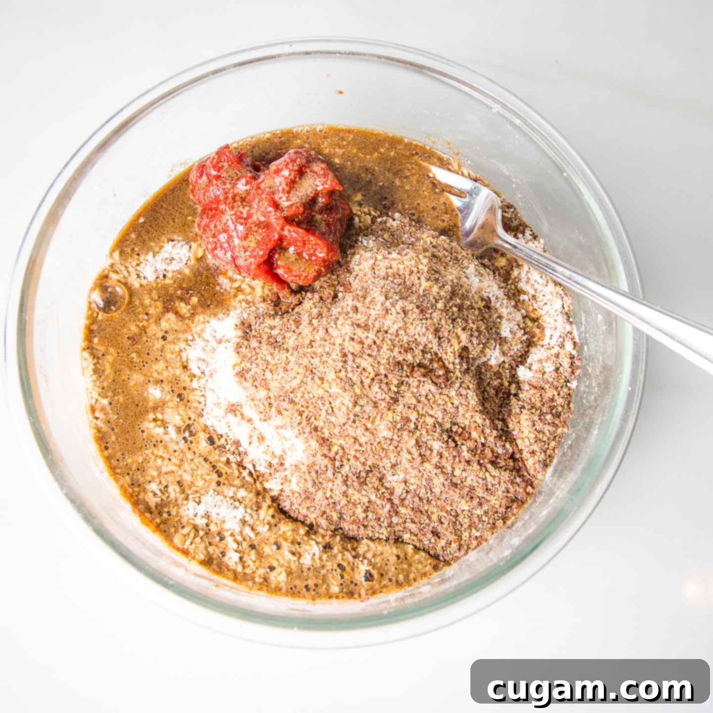 A glass bowl containing mashed beans, ground flax seeds, and a dollop of rich tomato paste, ready for mixing.