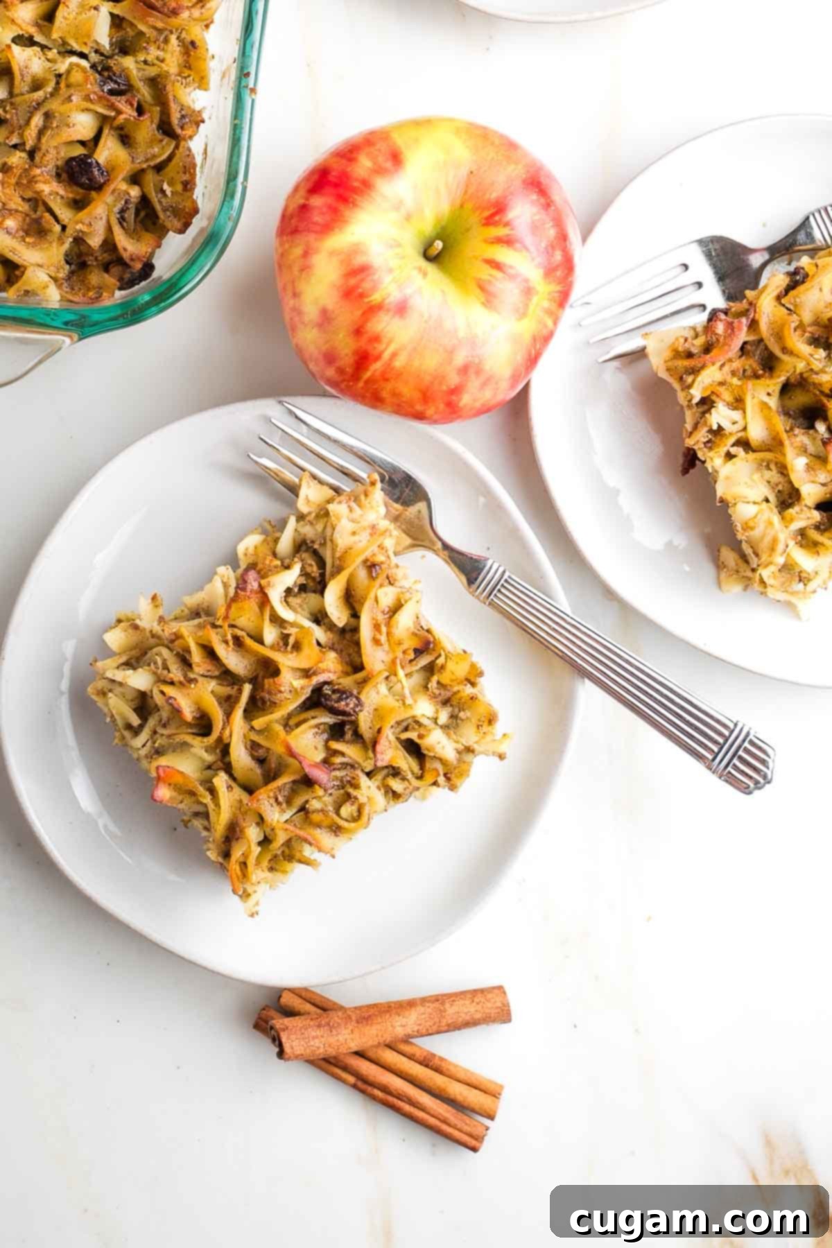 A perfectly portioned slice of golden-brown noodle kugel with tender raisins, set on a white plate. In the softly blurred background, a whole apple and fragrant cinnamon sticks hint at the dish's key ingredients.