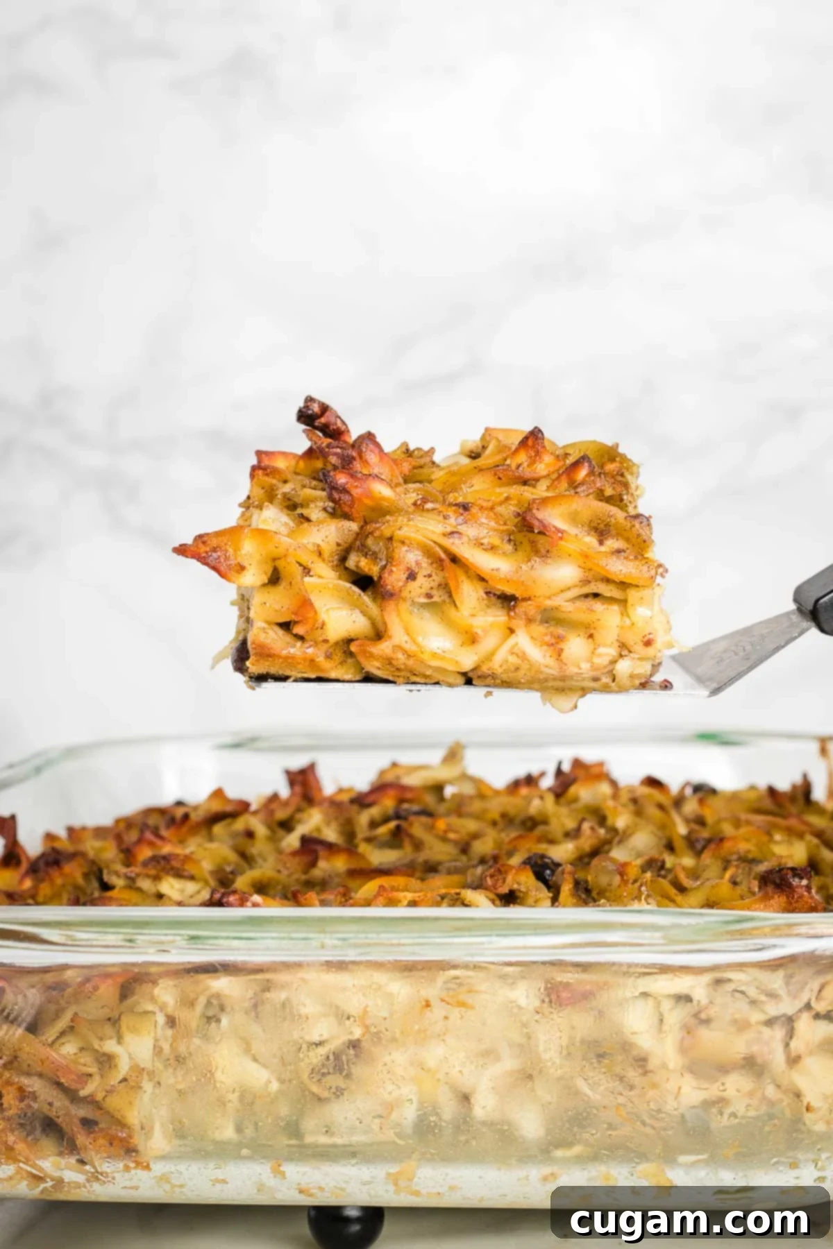 A glorious piece of baked noodle kugel, rich with apples and raisins, being gently lifted on a spatula above the rest of the casserole, ready to be served and enjoyed.