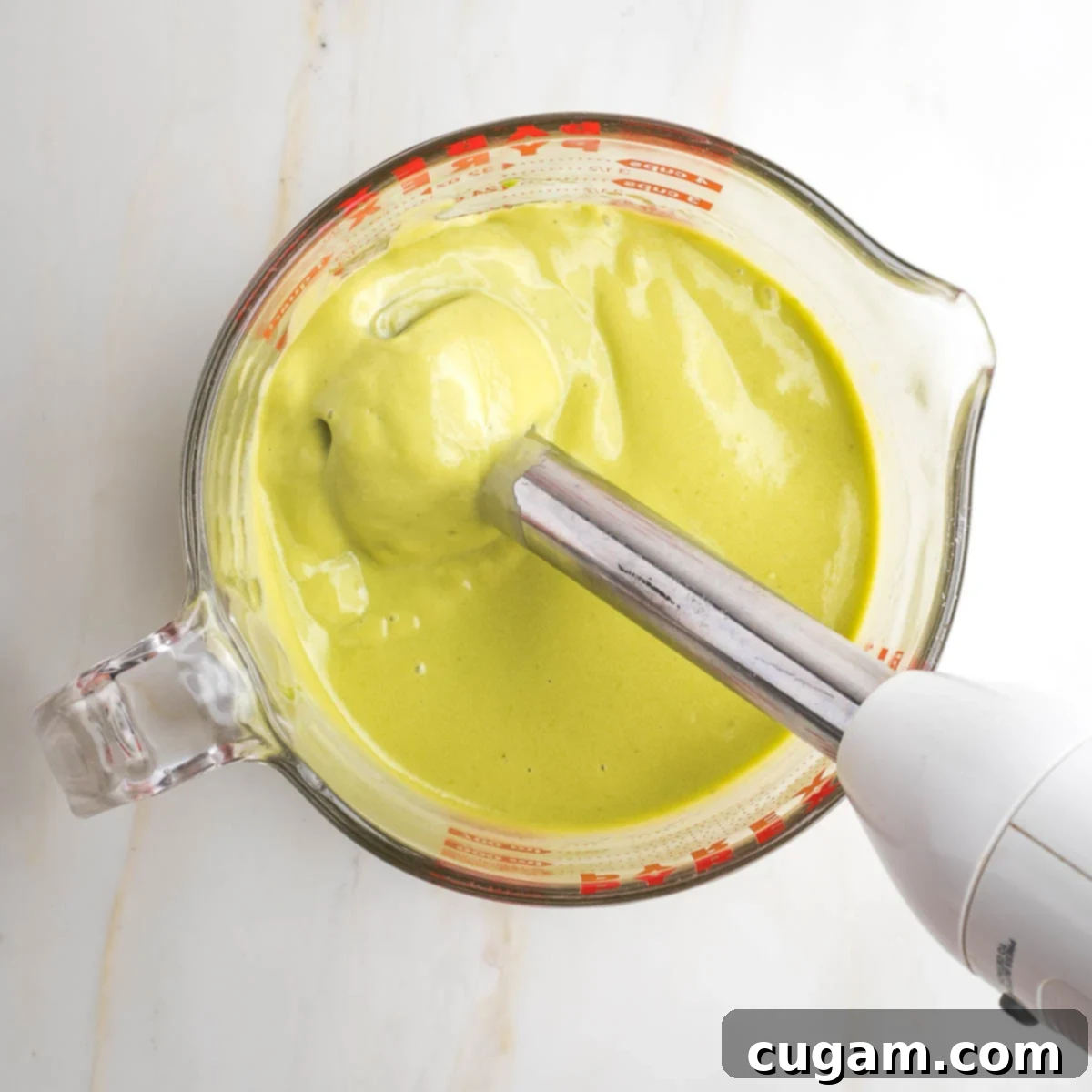 An immersion blender actively whirring in a large measuring cup, transforming the ingredients into a light green, velvety smooth, creamy mixture—the perfect custard consistency for the kugel.