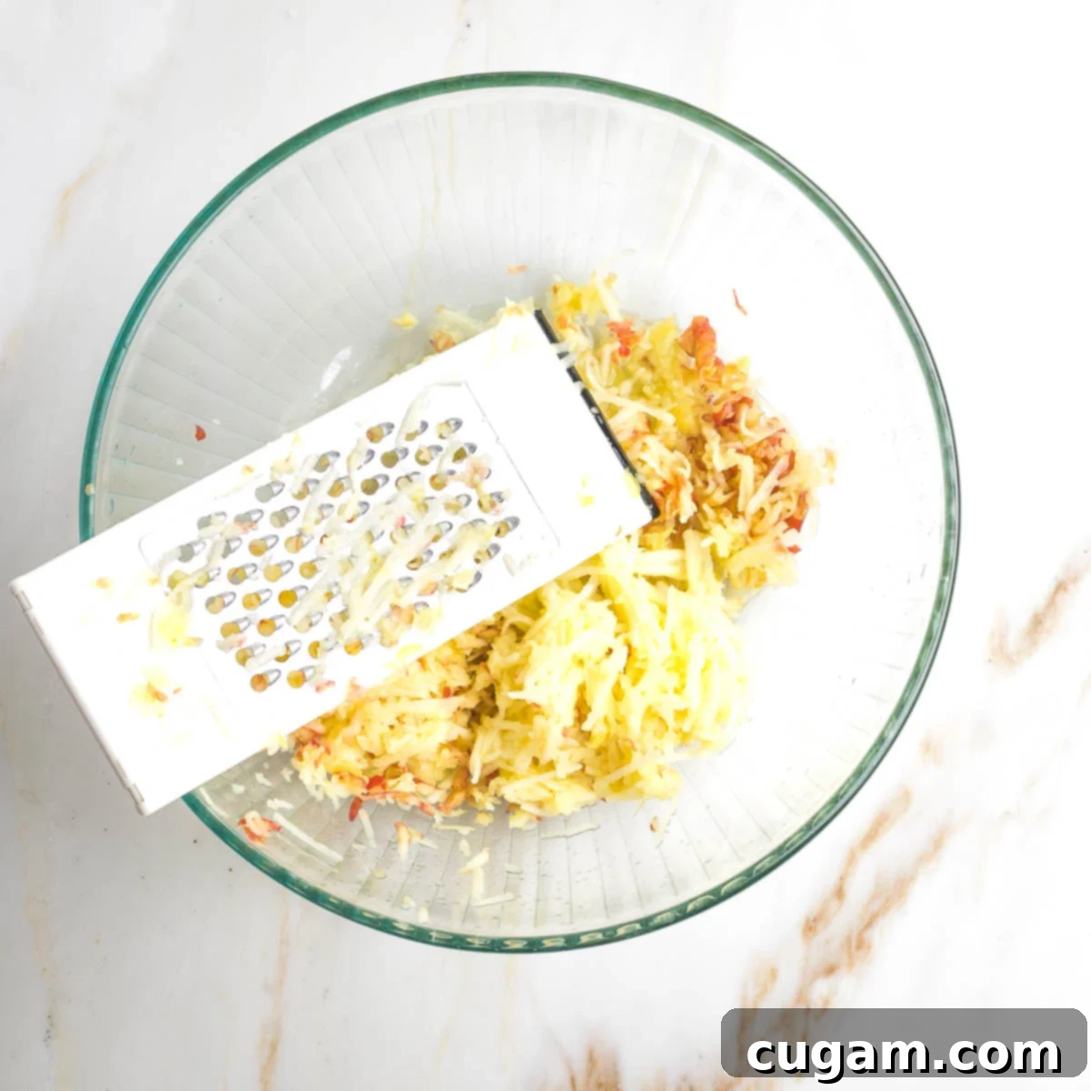 Hands are busy shredding fresh apples using a box grater, with the crisp, sweet strands falling into a large mixing bowl, ready to become part of the delicious kugel.
