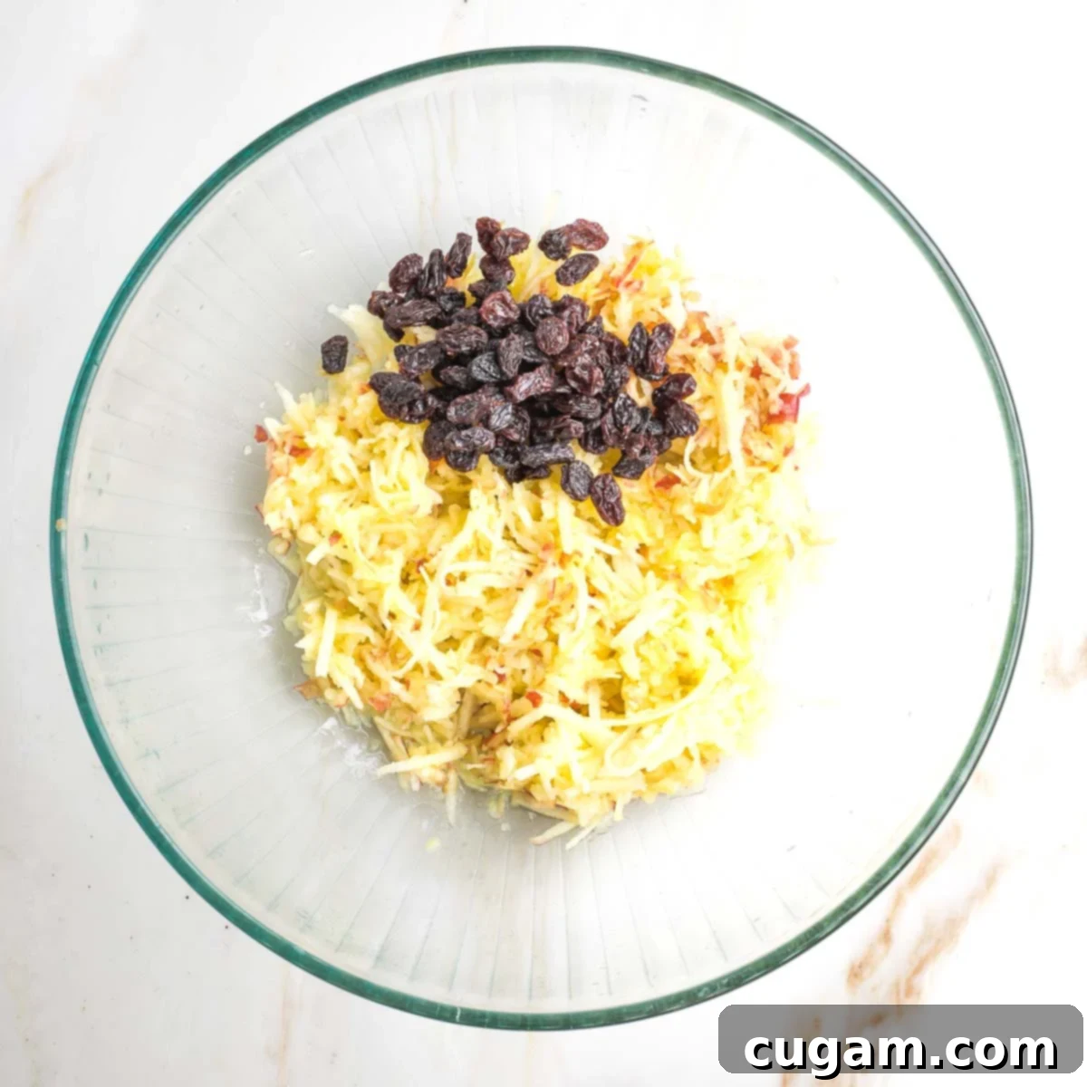 A clear glass mixing bowl holds a vibrant mix of freshly shredded apples and plump dark raisins, glistening slightly with natural apple juices, signaling moisture for the kugel.