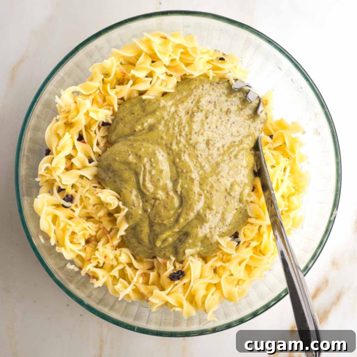 A large mixing bowl containing cooked noodles, to which a brownish, creamy pudding mixture is being poured and gently incorporated, illustrating the blending of the kugel's main components.