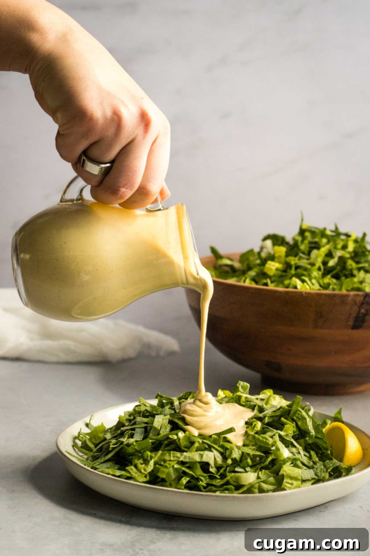 Creamy vegan tahini caesar dressing being generously poured over a fresh bed of sliced romaine lettuce on a white plate, with a bright yellow lemon half positioned elegantly nearby, hinting at the dressing's fresh citrus notes.