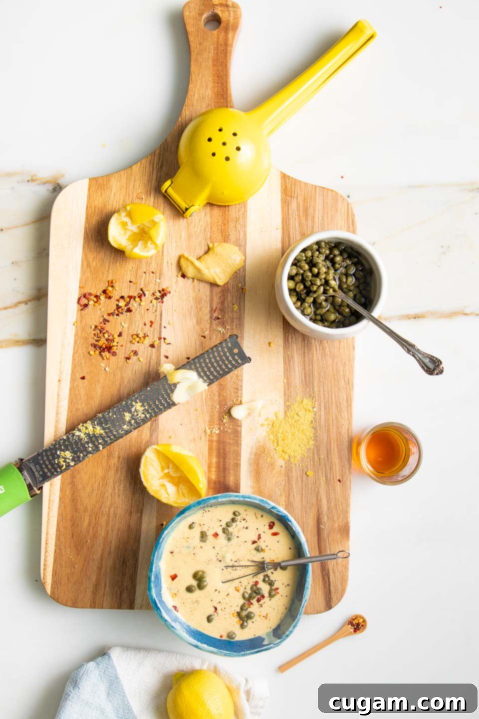 Close-up of fresh ingredients for a healthy tahini caesar dressing, including briny capers, a vibrant lemon cut in half, peeled garlic cloves, and a small pile of crushed red pepper flakes.