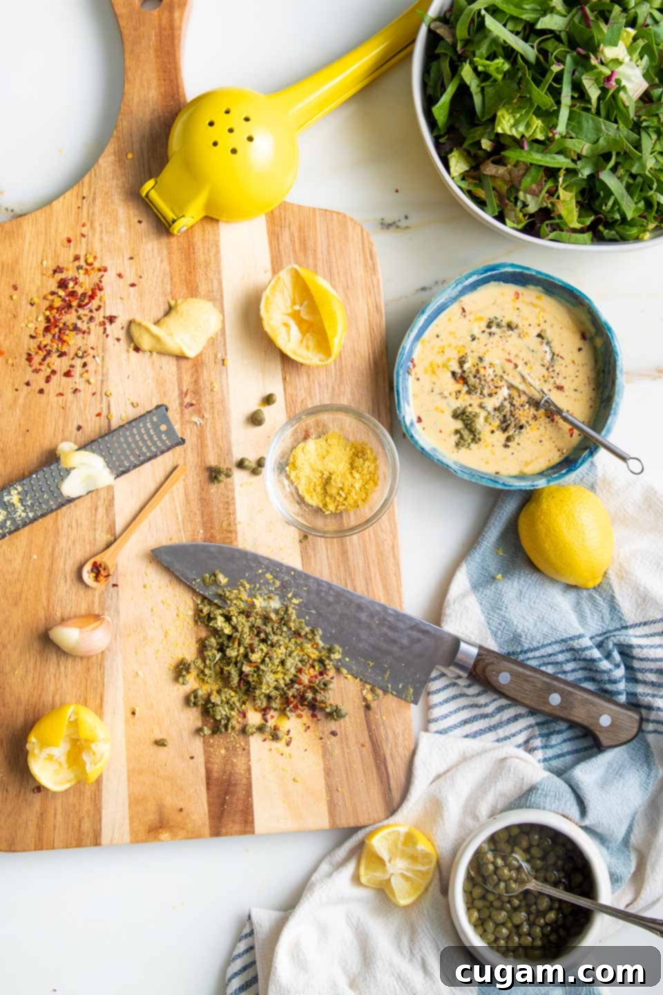 A bowl containing a whisked mixture of tahini, lemon juice, caper brine, and water, with finely chopped capers, zested garlic, and lemon zest waiting to be incorporated, for a hand-whisked Caesar dressing.