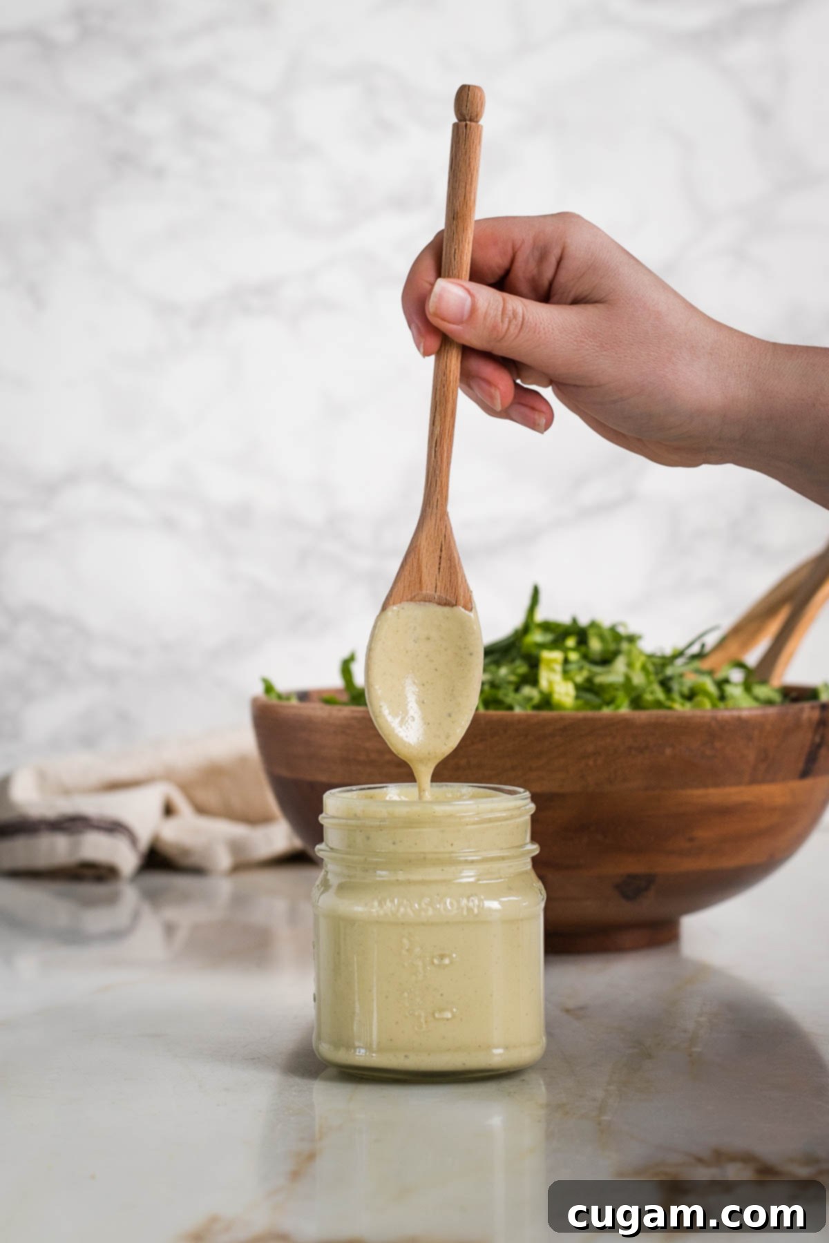 A wooden spoon dipping into a bowl of thick, creamy vegan tahini Caesar dressing, demonstrating its perfect consistency. In the background, a blurry bowl of fresh chopped greens awaits.