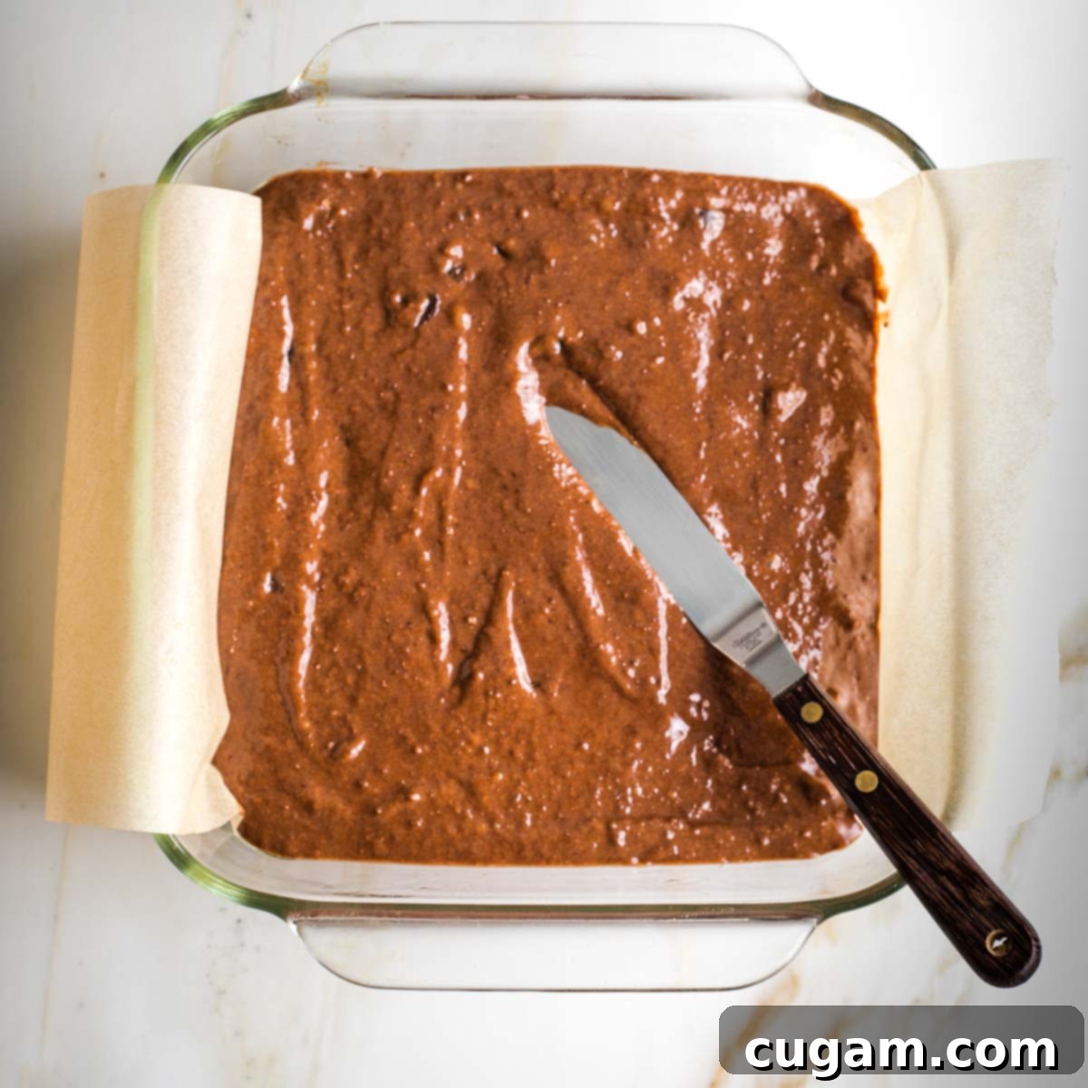 Brownie batter in a parchment-lined square glass pan, with an offset metal spatula spreading the batter evenly.