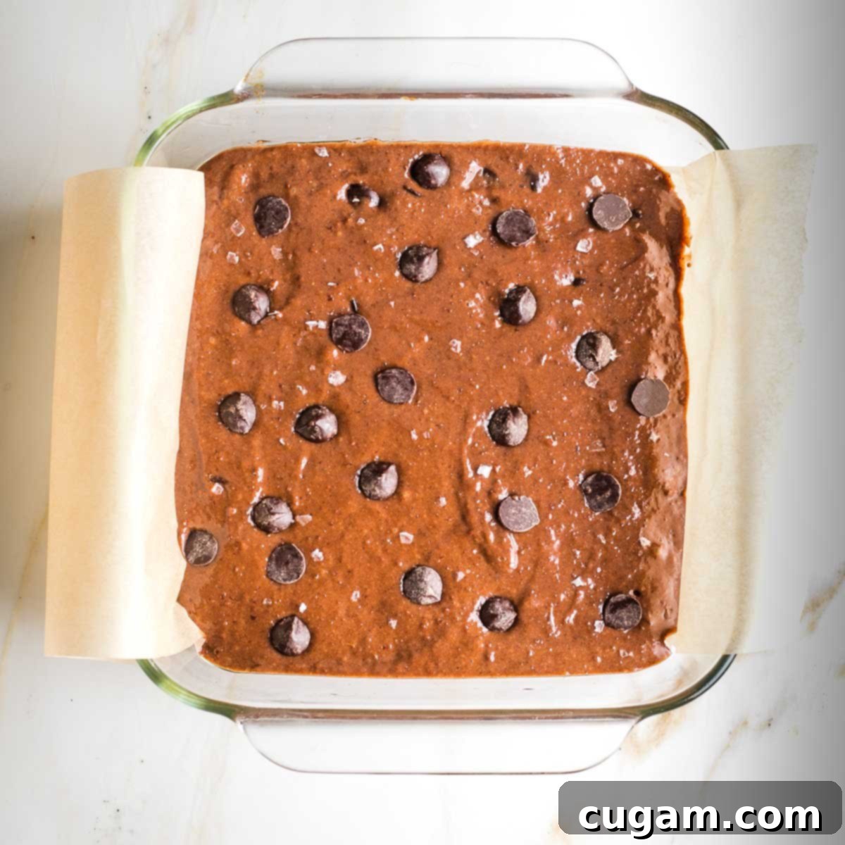 Vegan tahini brownie batter topped with an extra sprinkle of chocolate chips and flaky sea salt in a square glass pan, ready for baking.