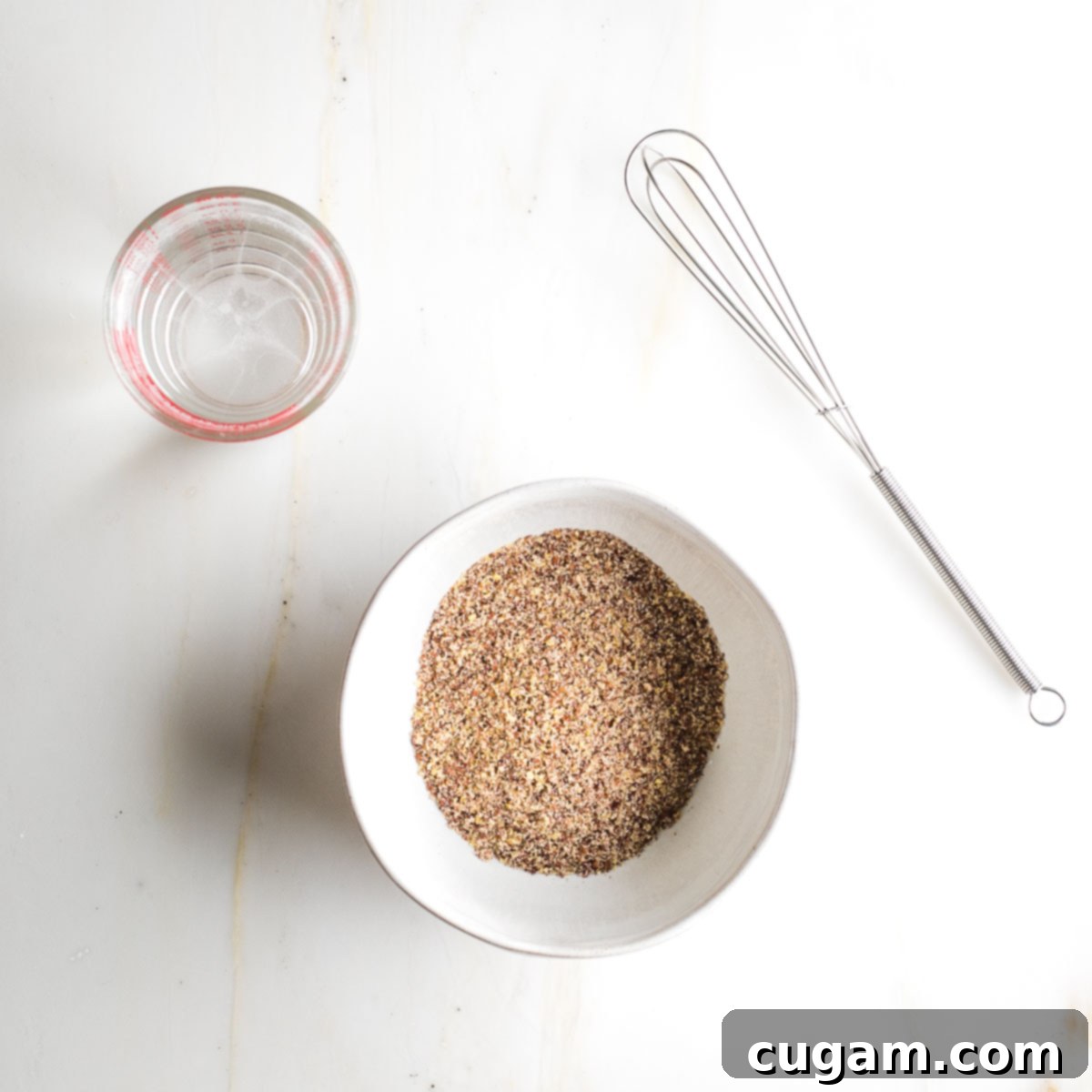 Small bowl with ground flax meal next to a glass of water and a small whisk, ready to make flax eggs.