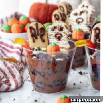 Clear cup of pudding and Oreos with gummy worms, pumpkin candy, sprinkles, headstone cookie, and mummy cookie, ready to be served.