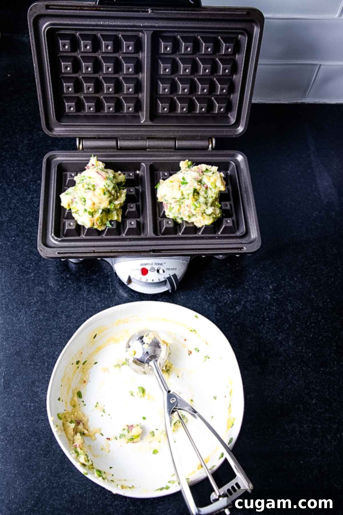 Waffle maker with large portions of potato waffle batter. Empty bowl with cookie scoop.