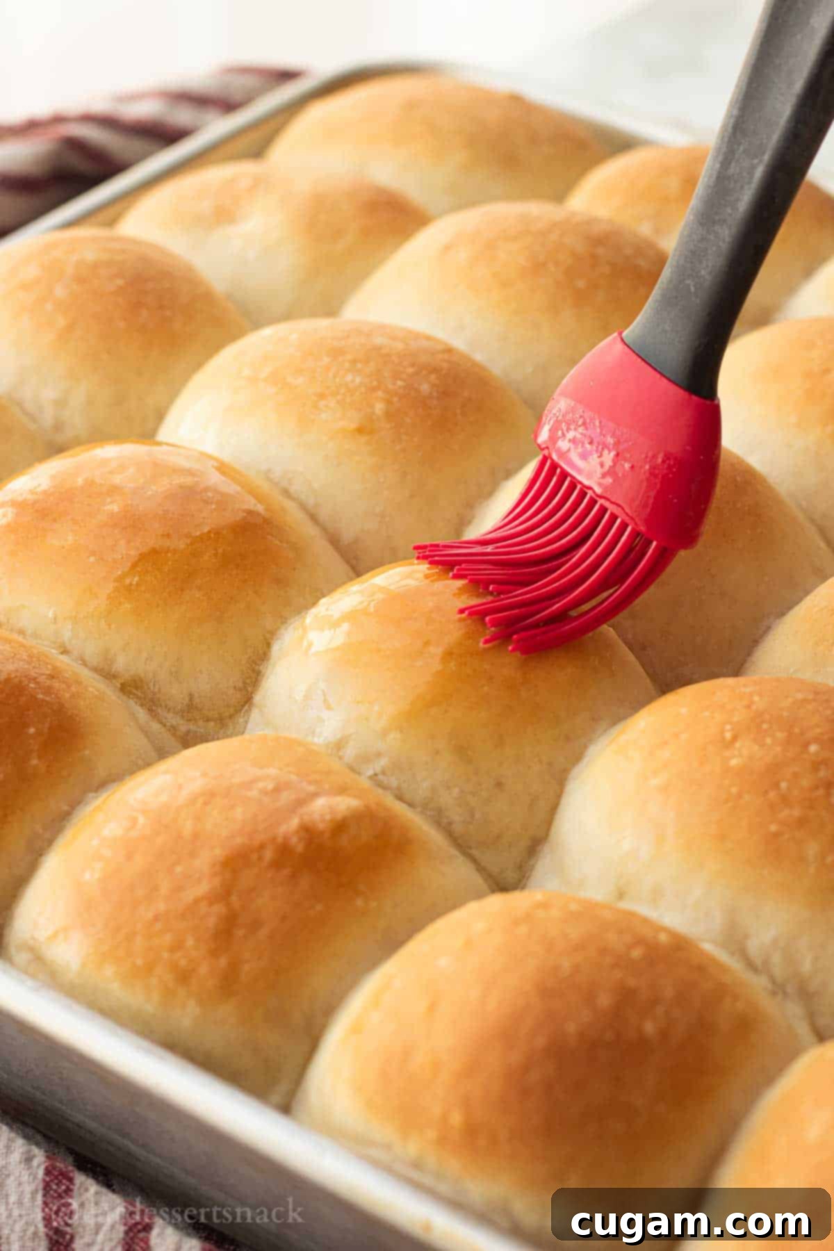 A vibrant red silicone brush generously applying melted butter to the tops of hot, freshly baked, golden fluffy dinner rolls still in the pan, creating a beautiful sheen.