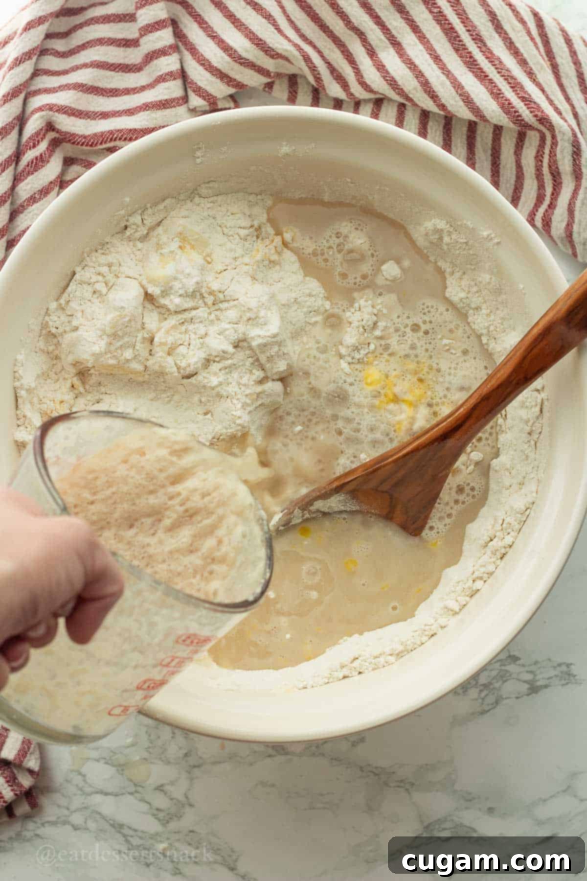 The activated yeast and honey mixture being carefully poured into a large white mixing bowl containing the dry ingredients like flour, butter, and eggs, ready for kneading.
