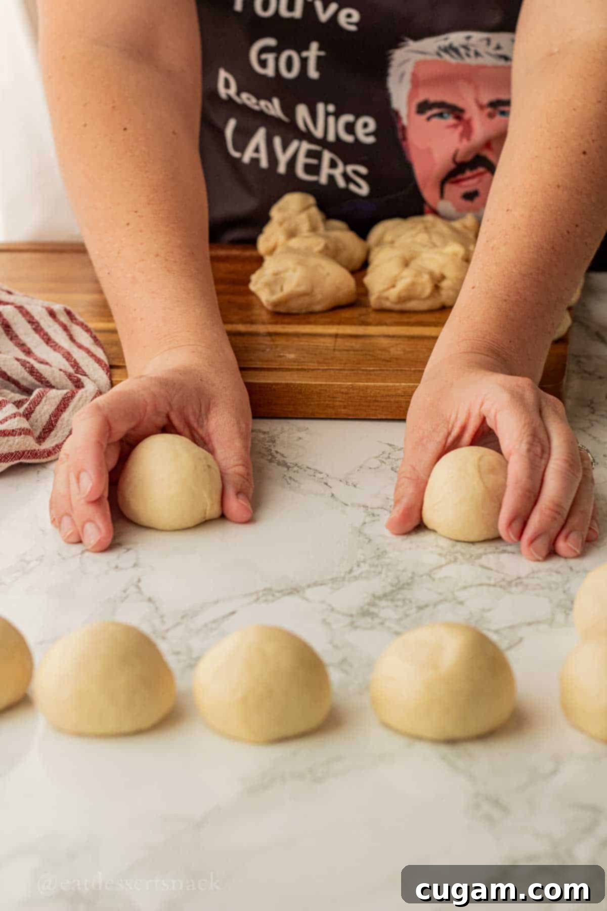 Skilled hands expertly rolling a portion of raw dinner roll dough into a smooth, perfectly round ball on a clean, flat marble surface, demonstrating the bakery-style technique.