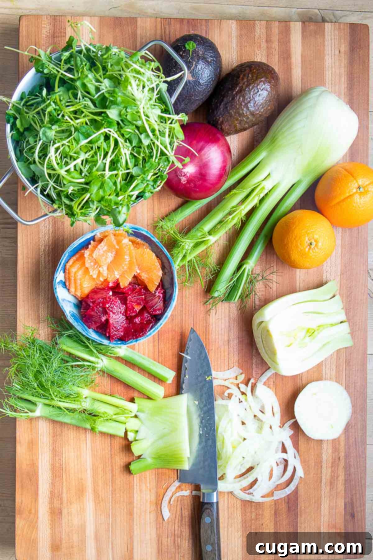 Knife cutting fennel into slices. Oranges, watercress avocado and onion in the background.