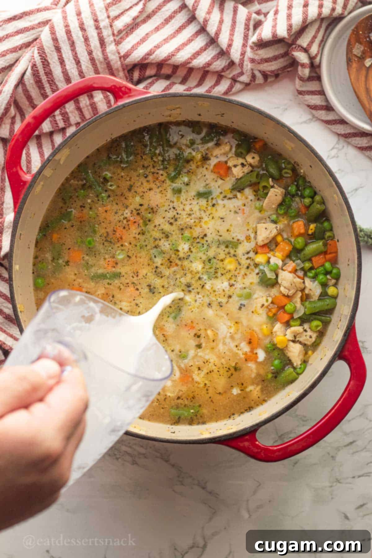 The milk and flour thickening mixture being slowly poured into a simmering red pot of chicken soup.