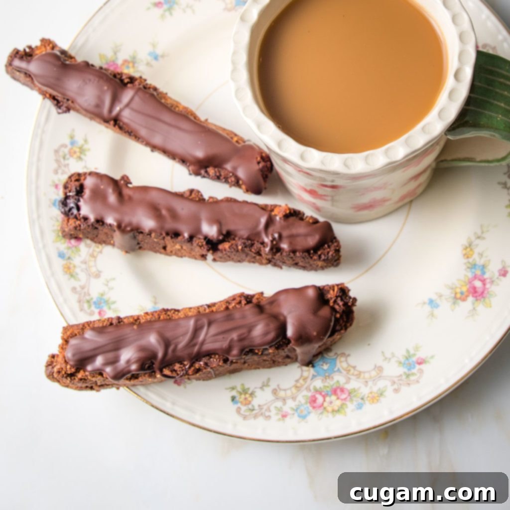 Three chocolate-covered vegan gluten-free biscotti on a flowery china plate next to a cup of coffee, ready for dunking.