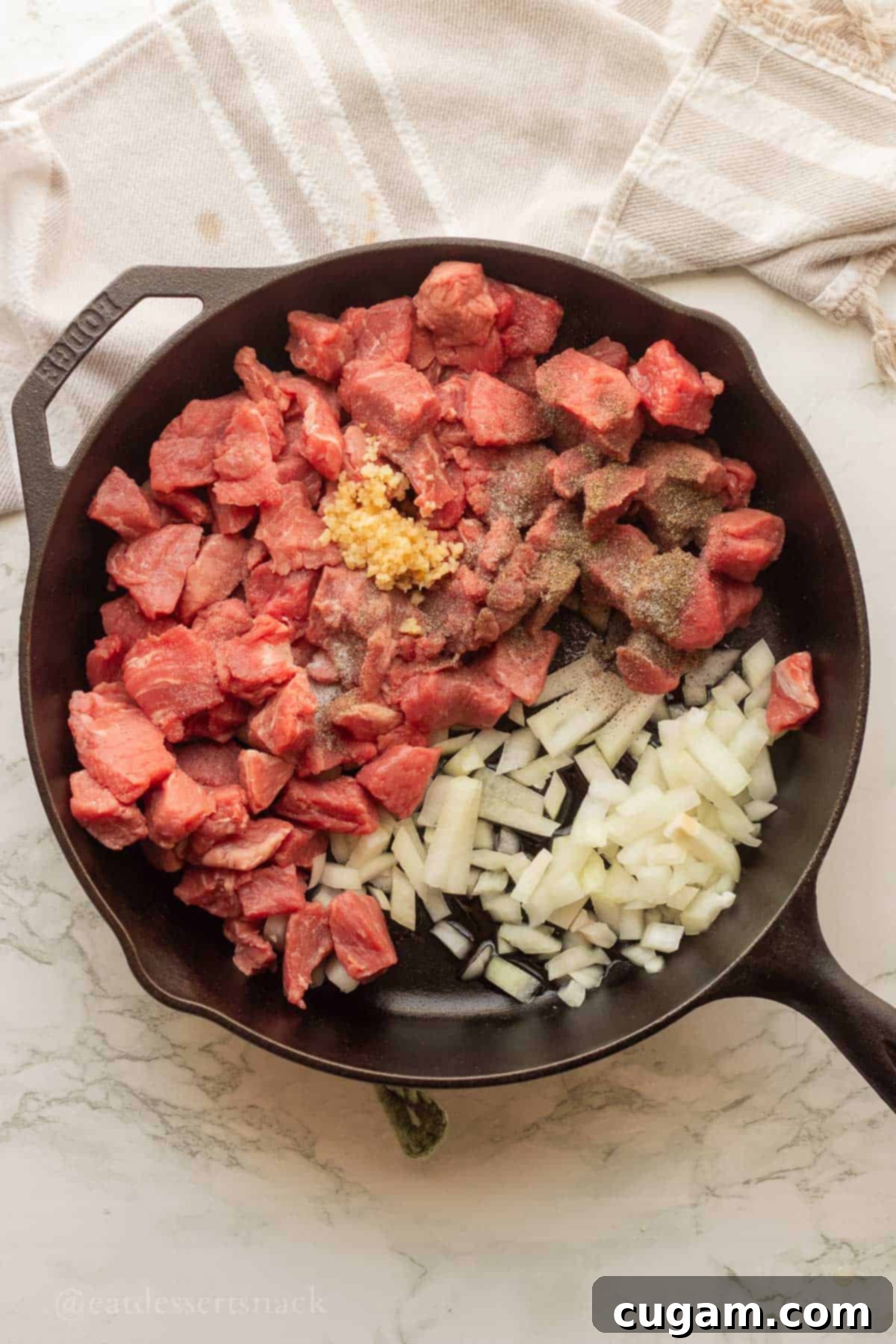 Raw beef chunks, garlic, and onion in black frying pan on marble.