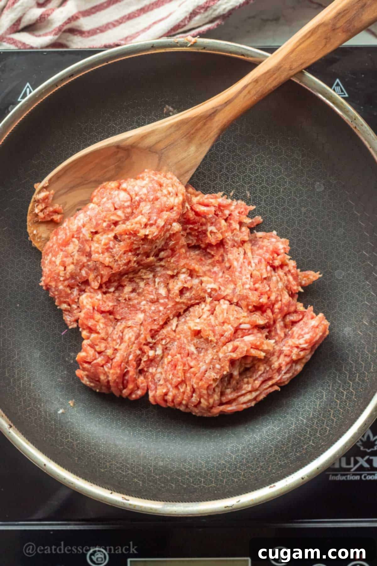 Raw ground beef being browned in a metal skillet with a wooden spoon, before being added to the soup.