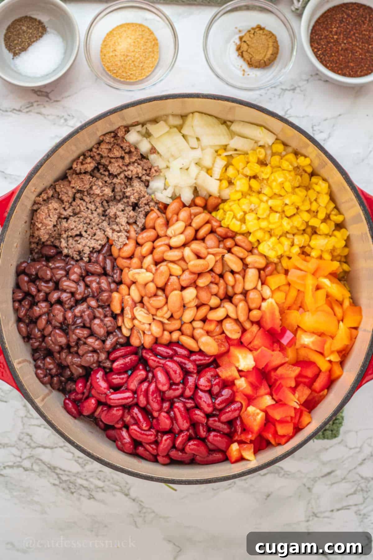 A vibrant mix of beans, cooked ground beef, diced onions, raw corn kernels, and colorful bell peppers combined in a red pot, ready for simmering on a marble countertop.