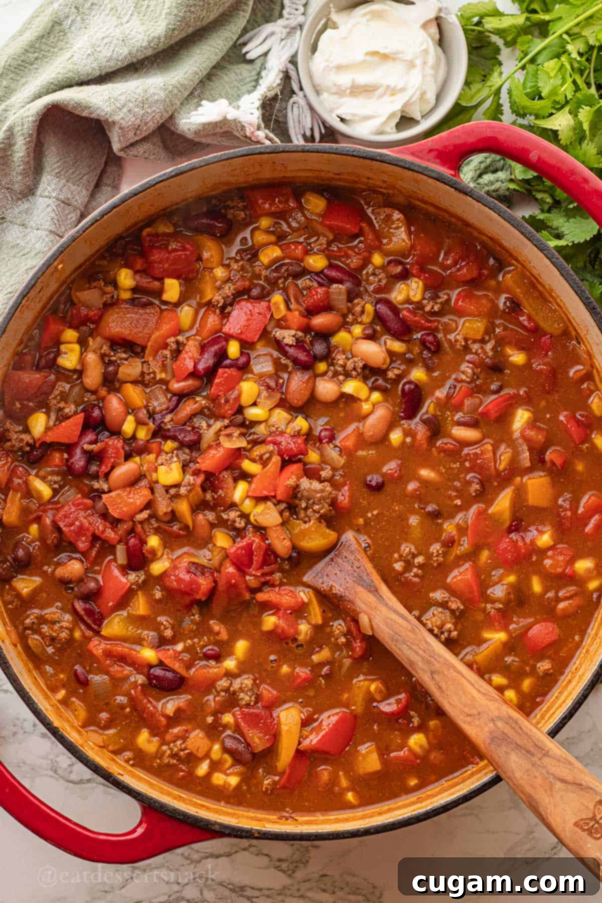 A red pot filled with richly colored, cooked taco soup, stirred with a wooden spoon, resting on a marble countertop, steam gently rising.