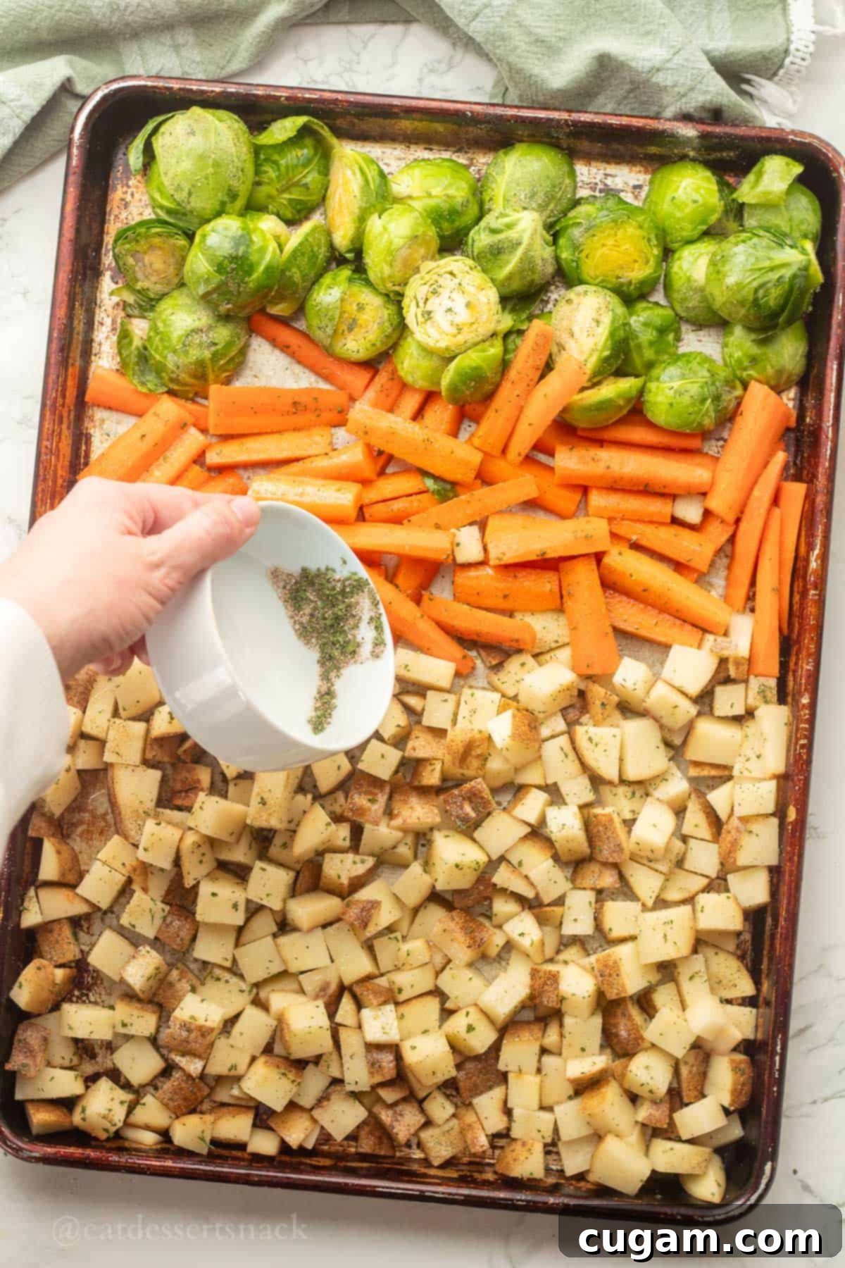 A hand sprinkling a savory seasoning blend evenly over raw vegetables (potatoes, carrots, Brussels sprouts) on a metal baking pan.
