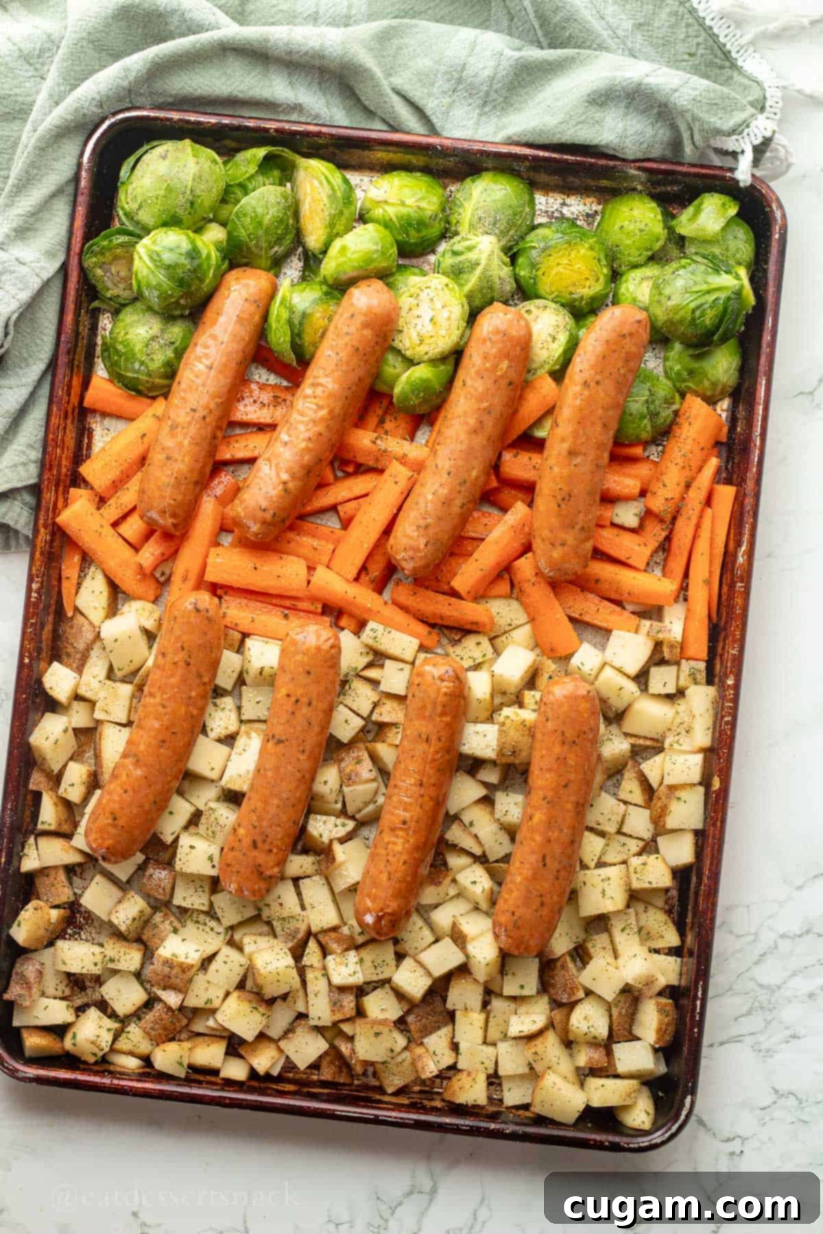 Raw chicken sausages neatly arranged on top of a bed of seasoned potatoes, Brussels sprouts, and carrots on a metal baking pan, ready for the oven.