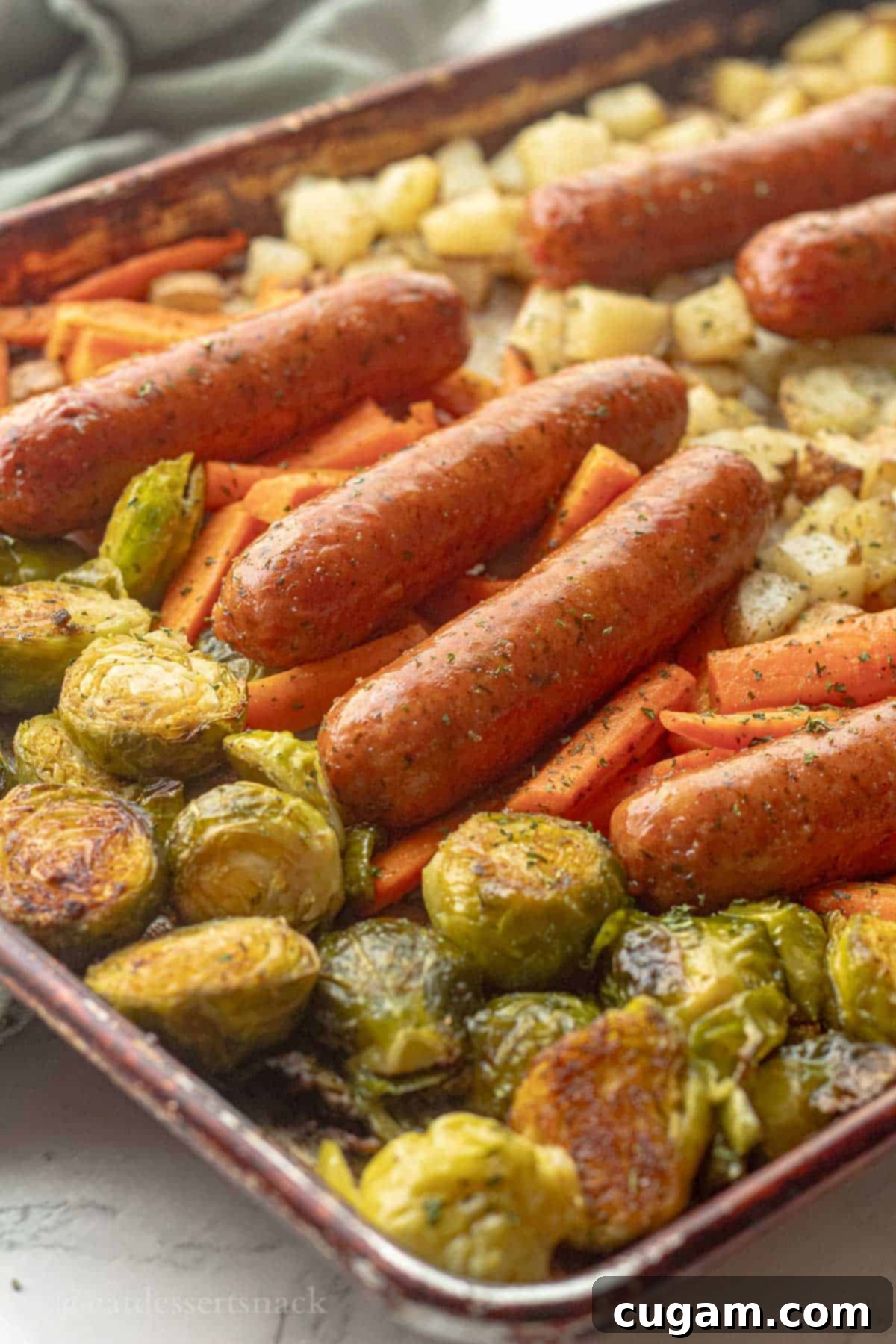 A close-up shot of perfectly cooked chicken sausage and golden-brown roasted vegetables, including potatoes, carrots, and Brussels sprouts, served directly on the metal baking pan.