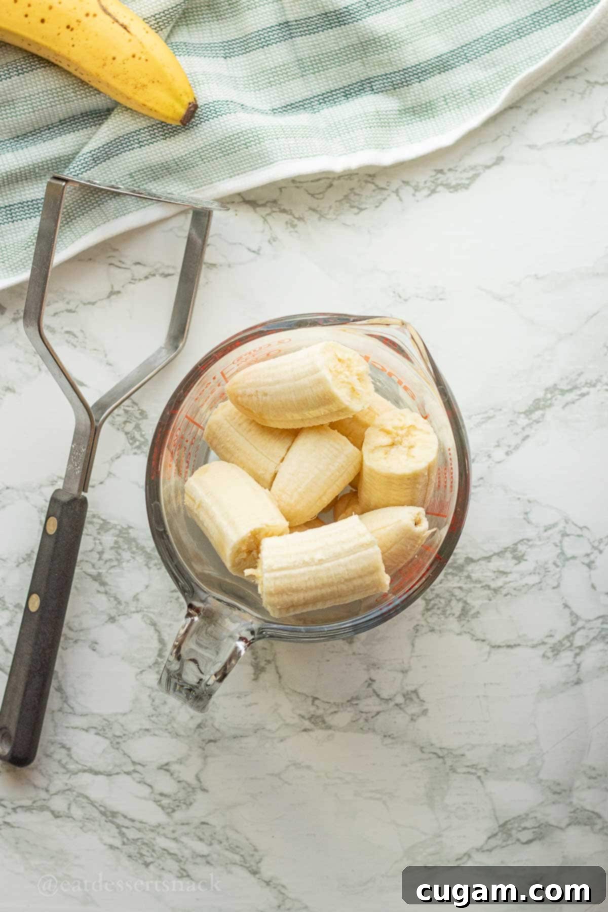 Bananas in glass measuring cup with potato masher on marble countertop.