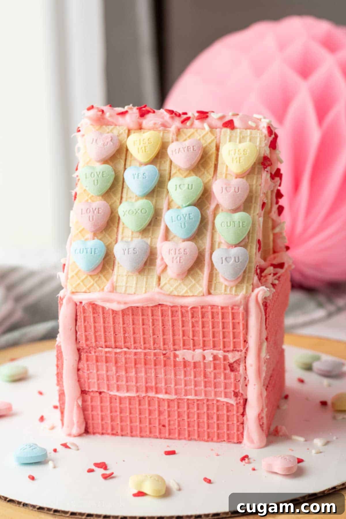 Pink wafer cookie house with Valentine candy on roof on white plate.