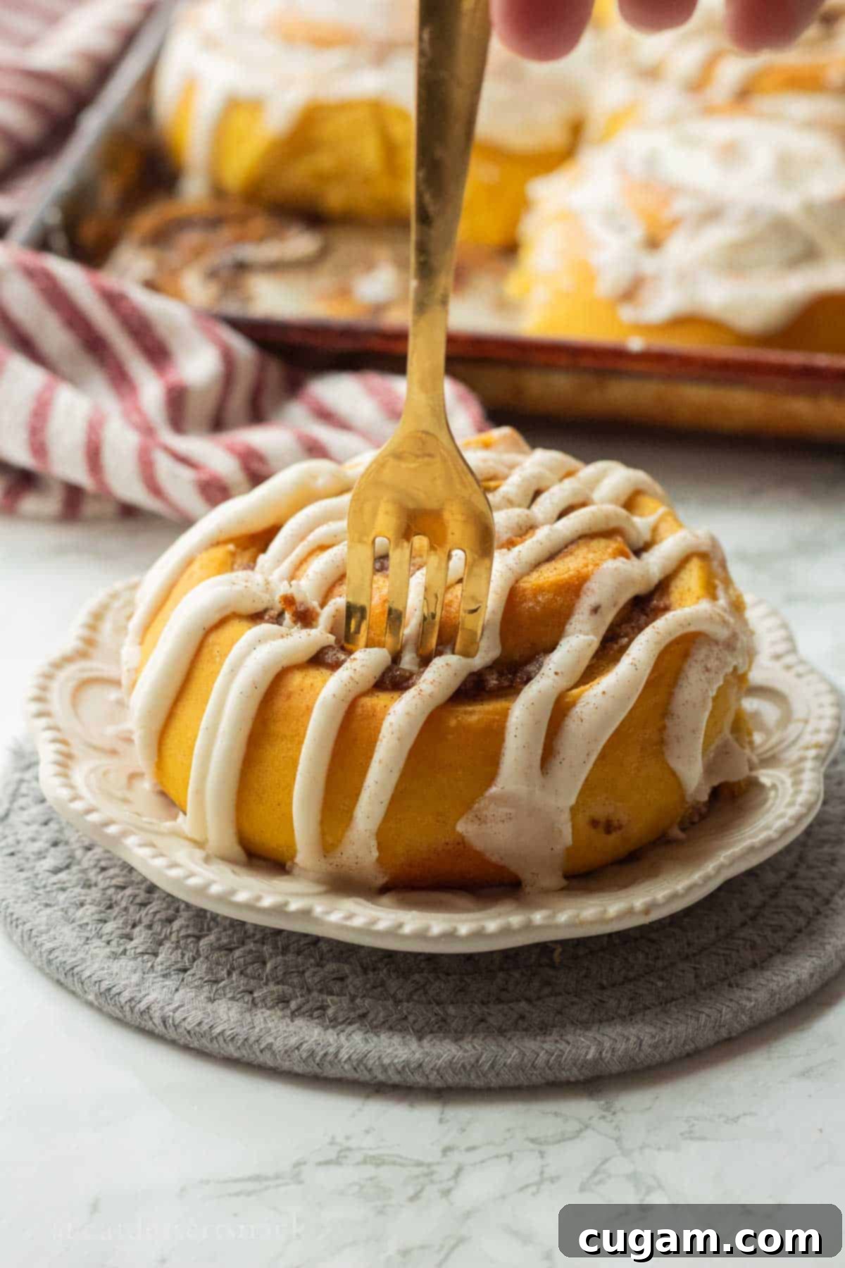 Gold fork in iced pumpkin cinnamon roll on white plate, ready to eat.