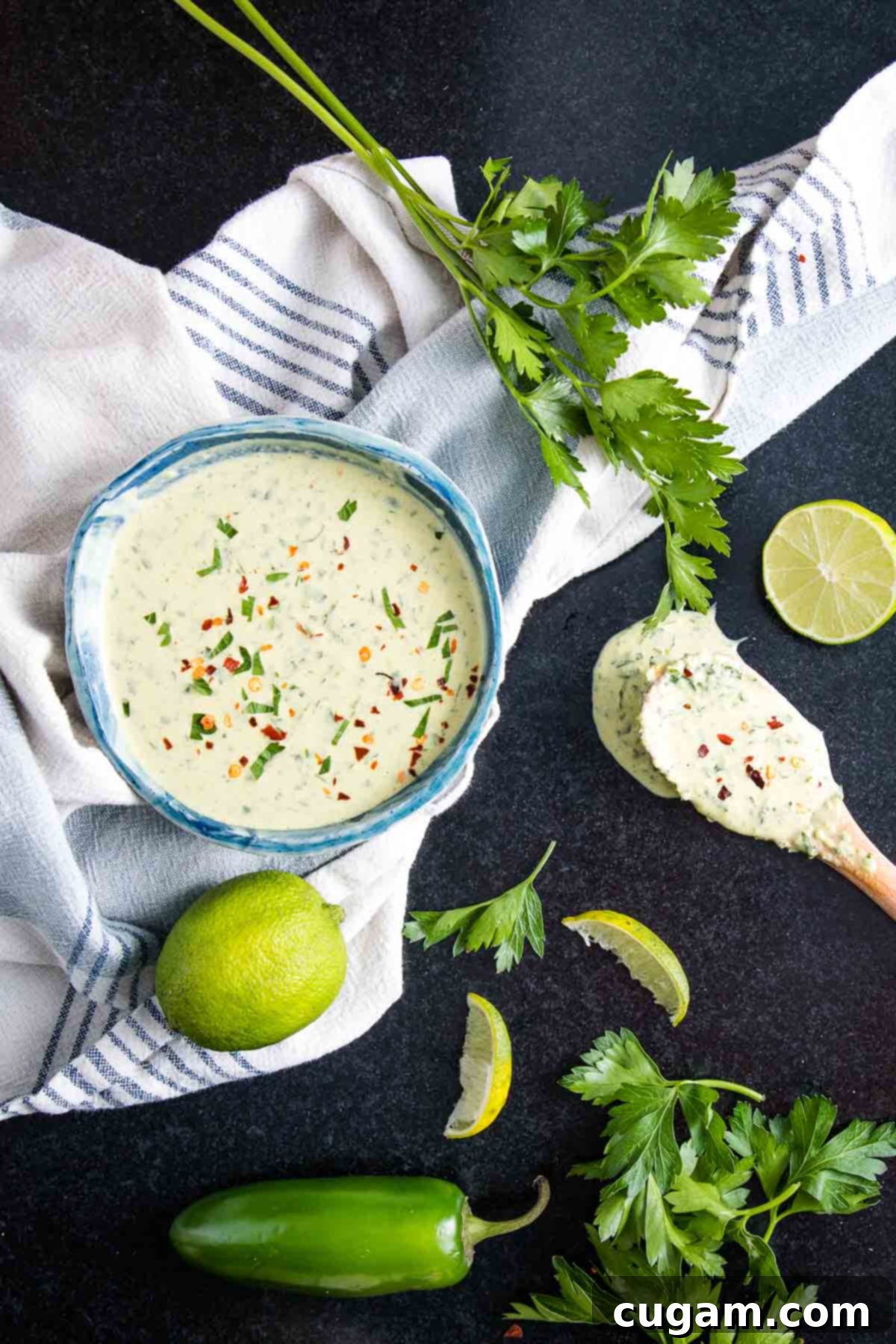 Green tahini sauce in a white bowl, elegantly surrounded by fresh limes, sprigs of vibrant parsley, and whole green jalapenos, highlighting its key ingredients.
