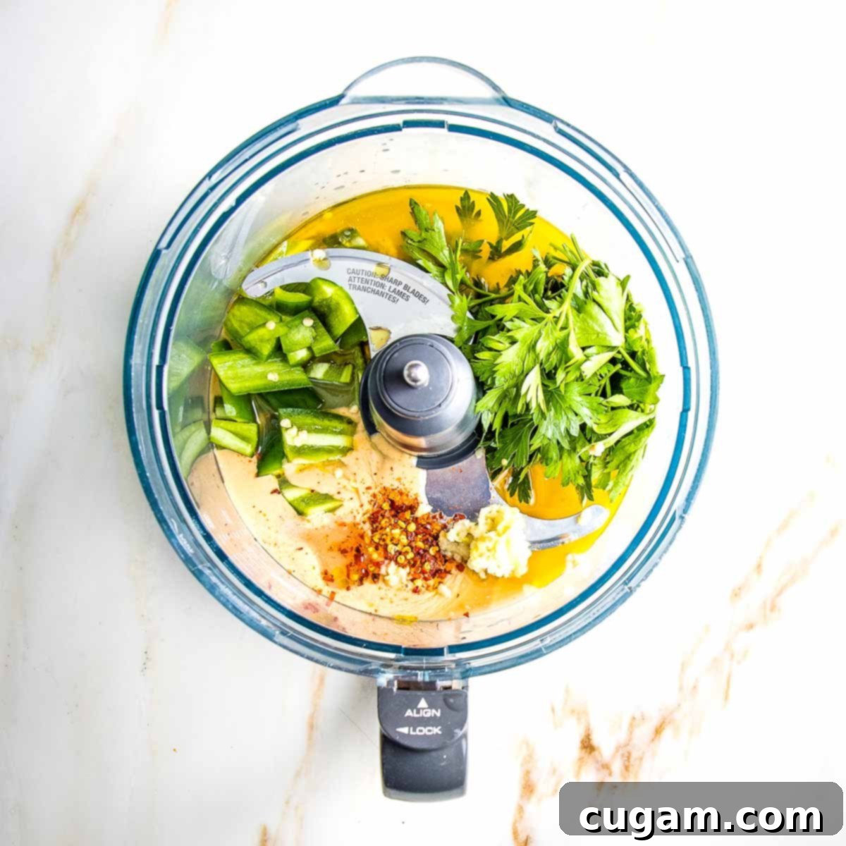 A food processor bowl containing the initial ingredients for green tahini sauce: creamy tahini, chopped jalapeno, fresh parsley, crushed red pepper, and grated garlic, ready for blending.