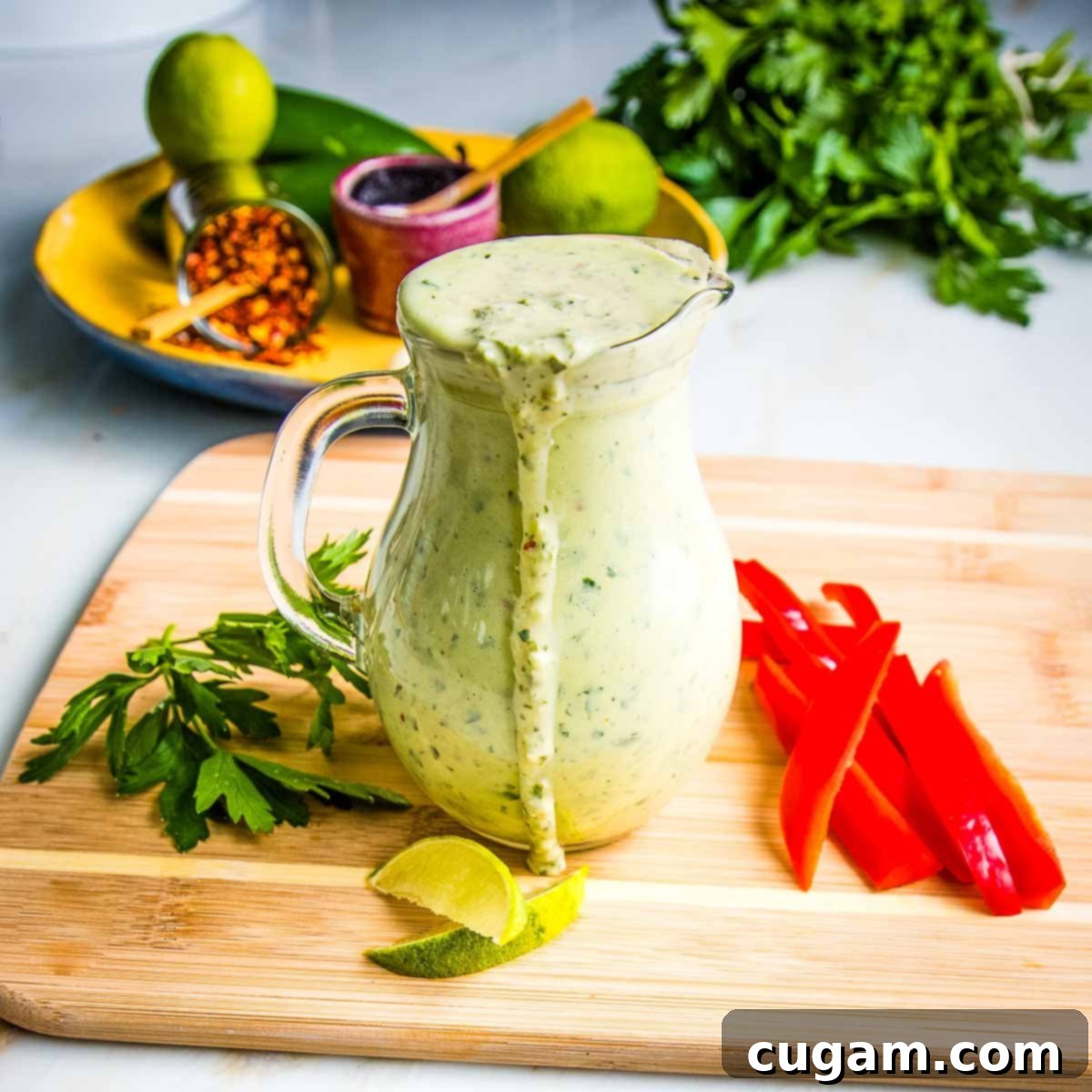 A clear pitcher of green tahini sauce with some sauce dripping over the edge onto a wooden board, surrounded by an assortment of colorful cut-up vegetables.