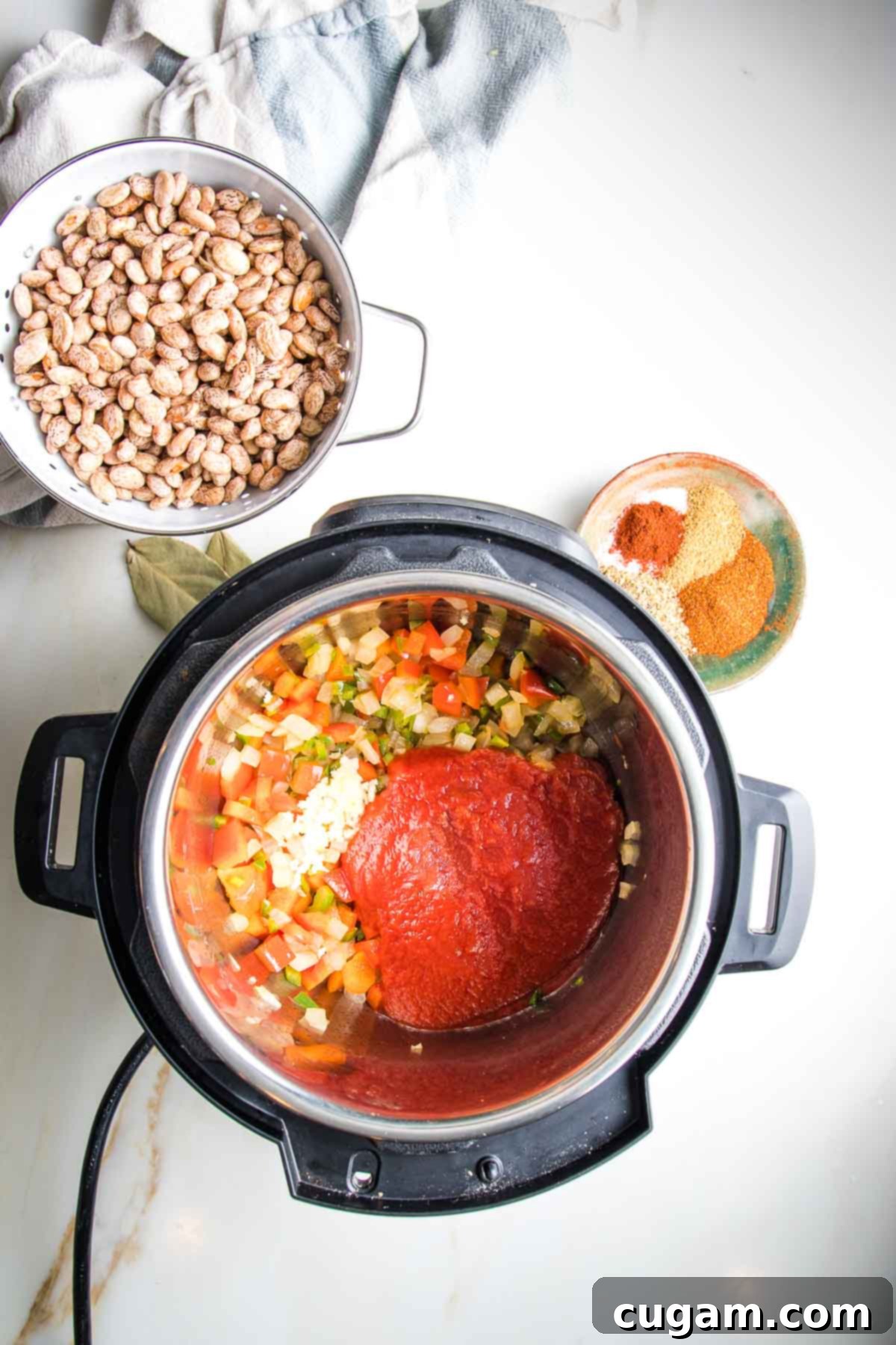 Tomato sauce poured on top of sauteed veggies in an instant pot, plus a dish of spices and a bowl with soaking pinto beans.